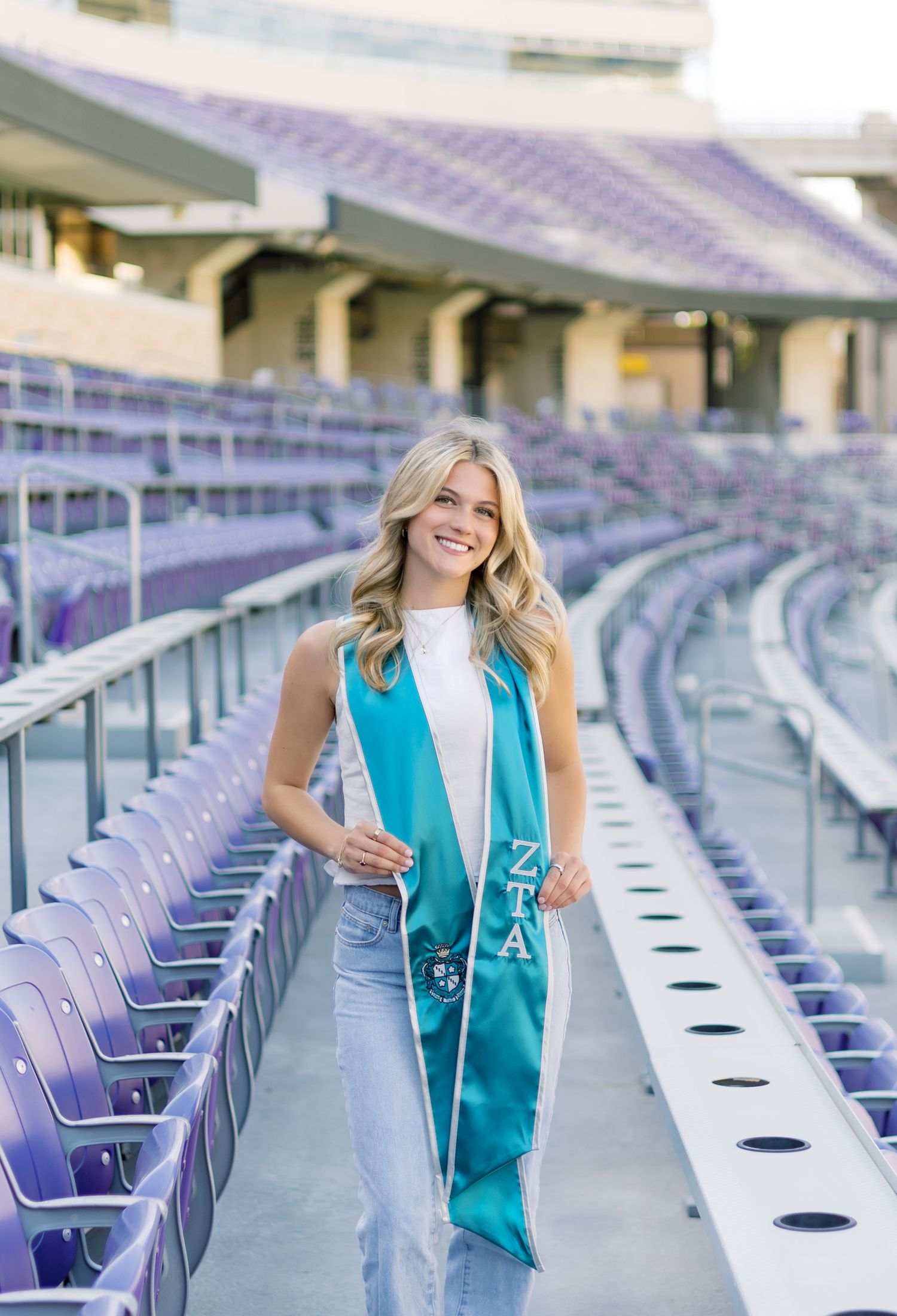 Smiling blonde woman wearing teal Zeta Tau Alpha graduation stole in purple TCU stadium stands.
