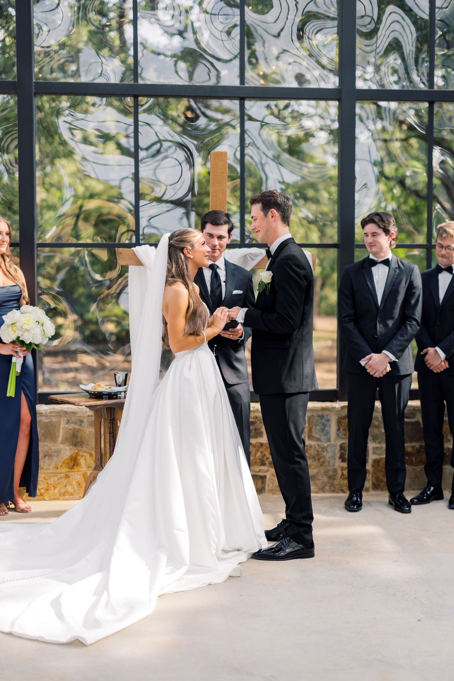 Bride and groom exchange vows at elegant outdoor wedding ceremony with groomsmen and stained glass backdrop.