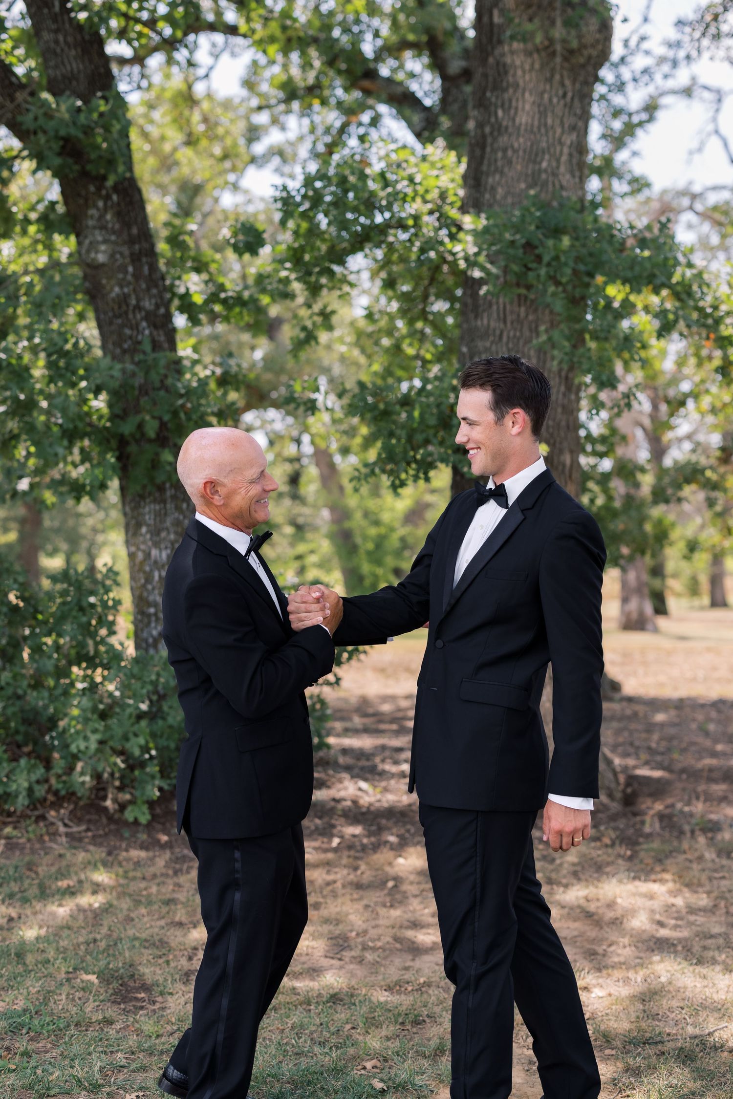 Two men in black tuxedos shake hands outdoors among trees, likely at a wedding celebration.
