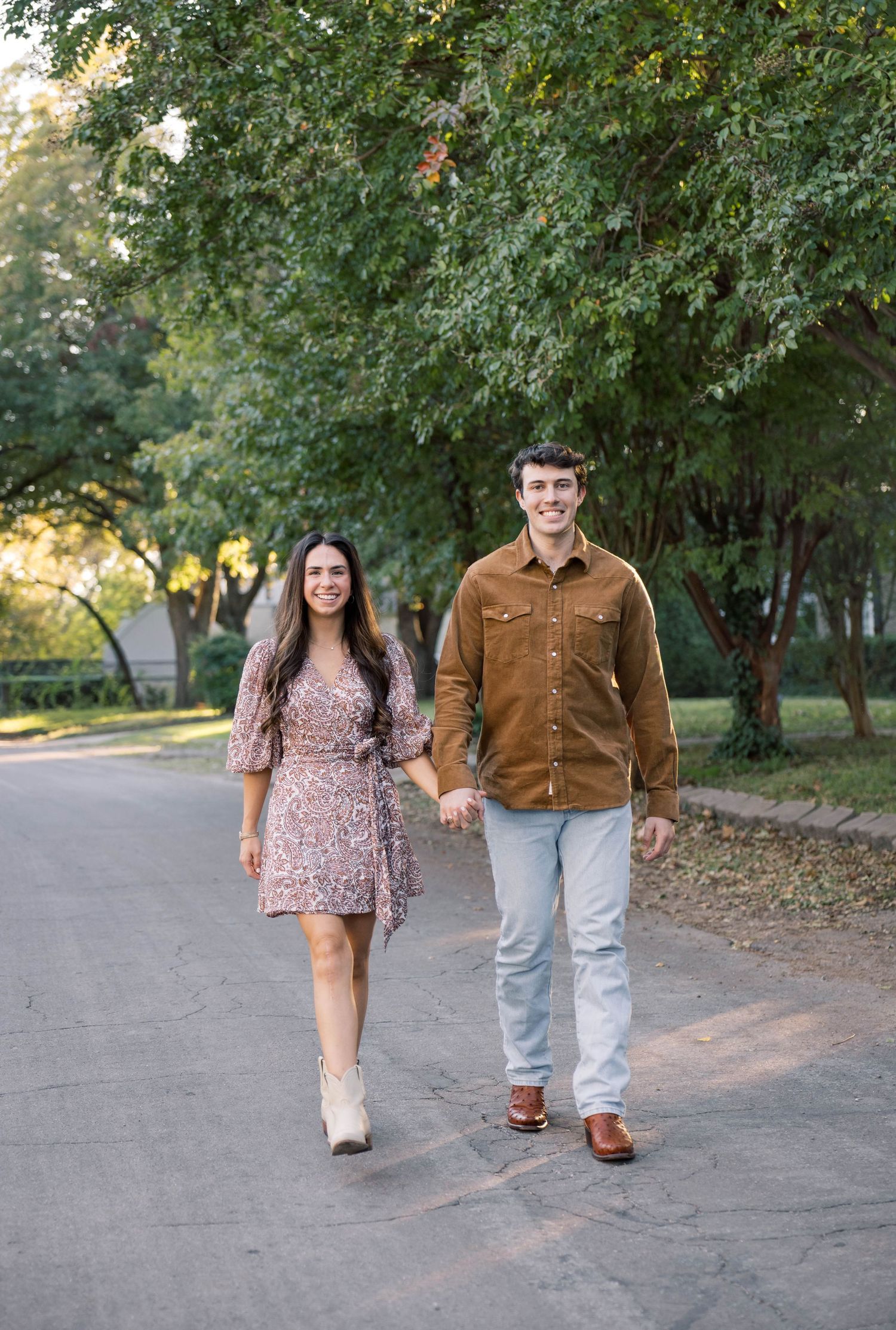 Smiling couple walking hand-in-hand on a shaded path, wearing coordinated brown and floral autumn outfits.