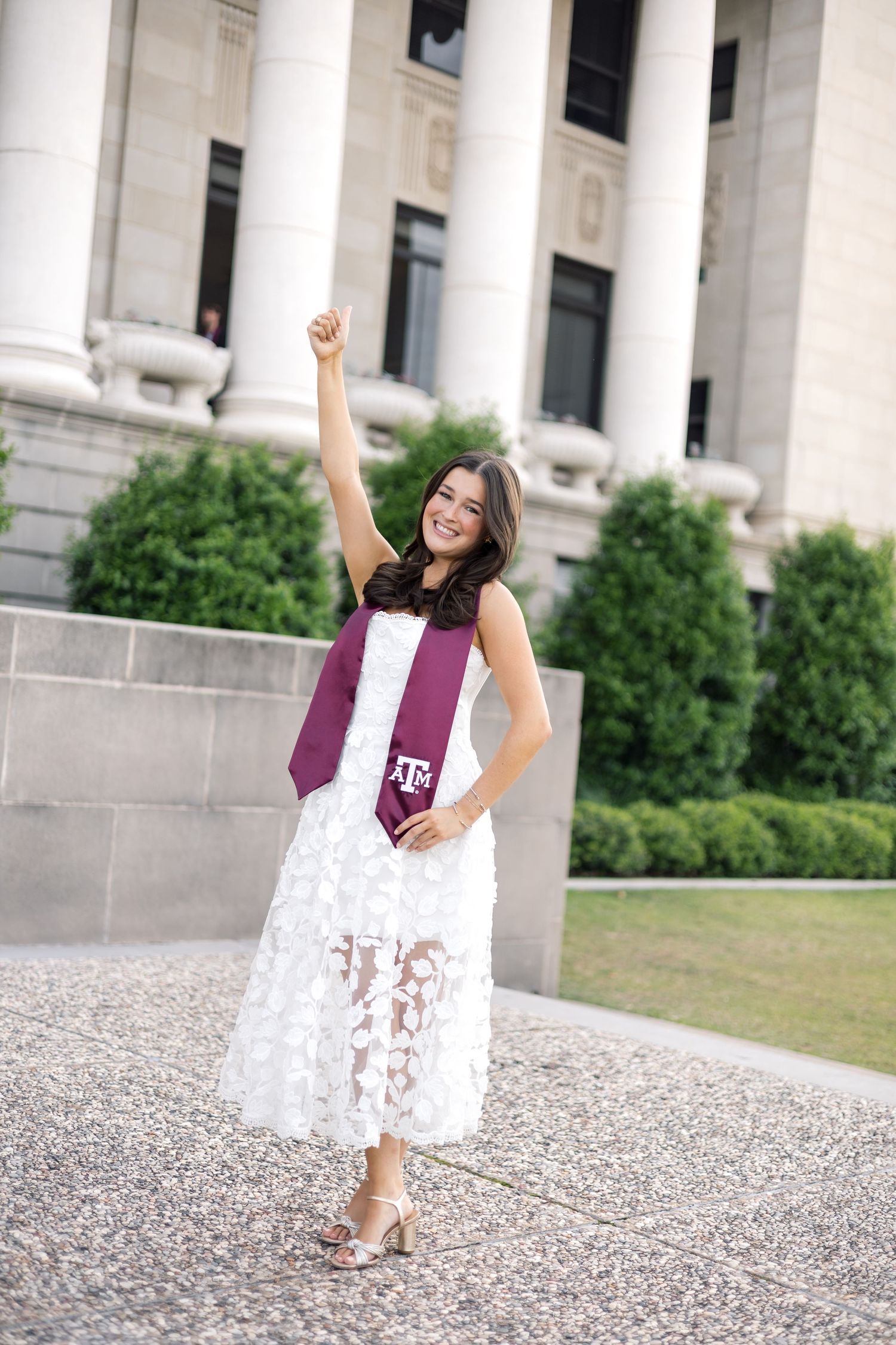 Excited Texas A&M graduate in white lace dress with maroon stole raising fist in celebration.