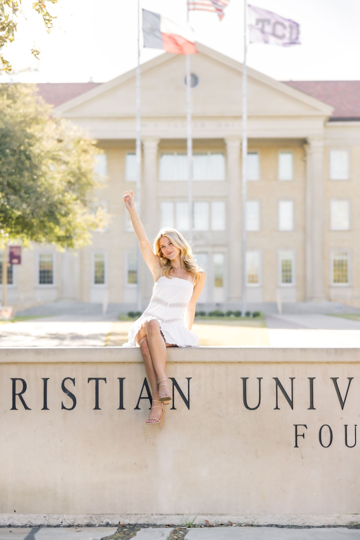 Blonde woman in white dress sitting on TCU Christian University Foundation wall with arm raised.