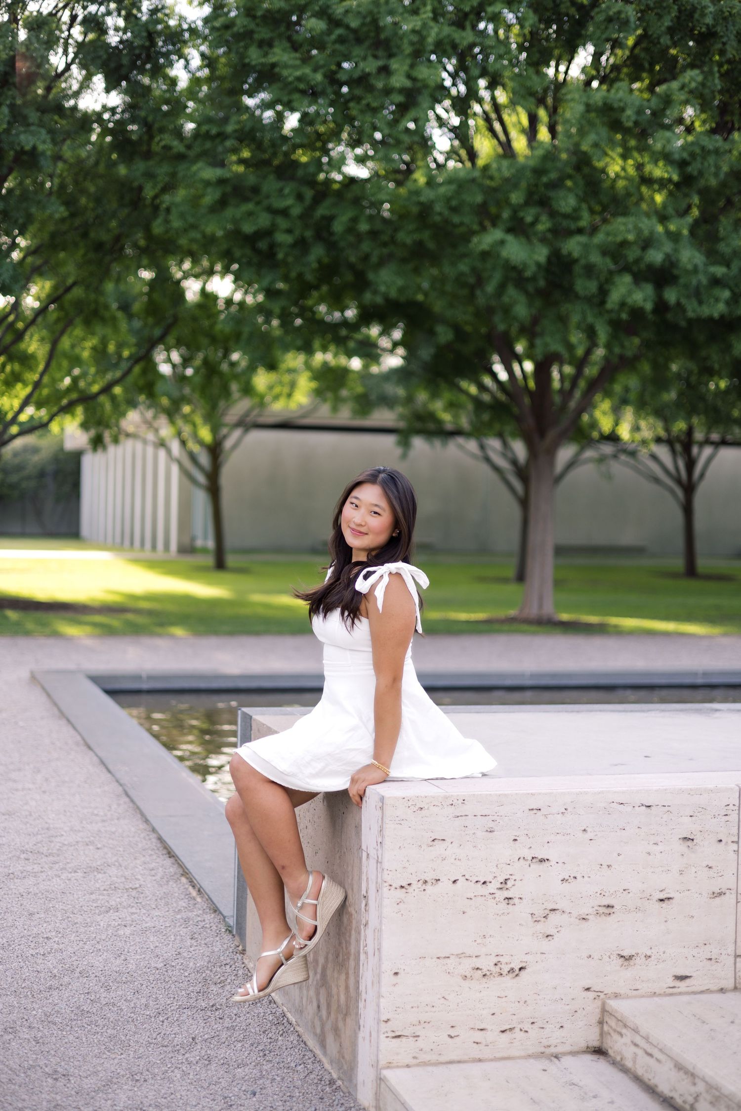 Young woman in white dress sitting by a reflecting pool in a lush green park, smiling gracefully.
