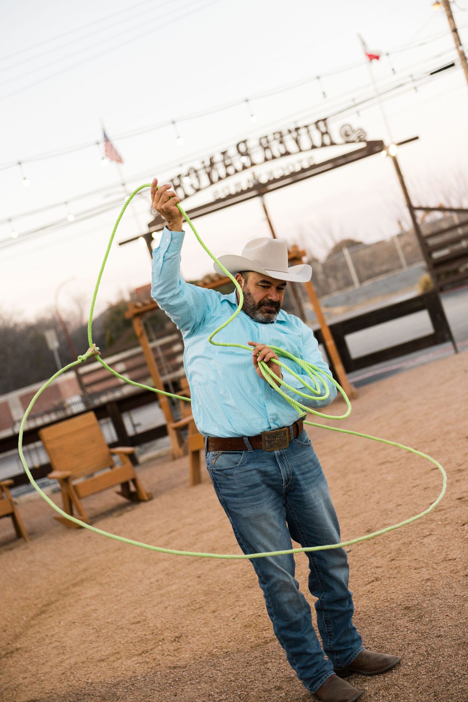 A cowboy in a white hat skillfully spins a bright green lasso rope outdoors at a western ranch.