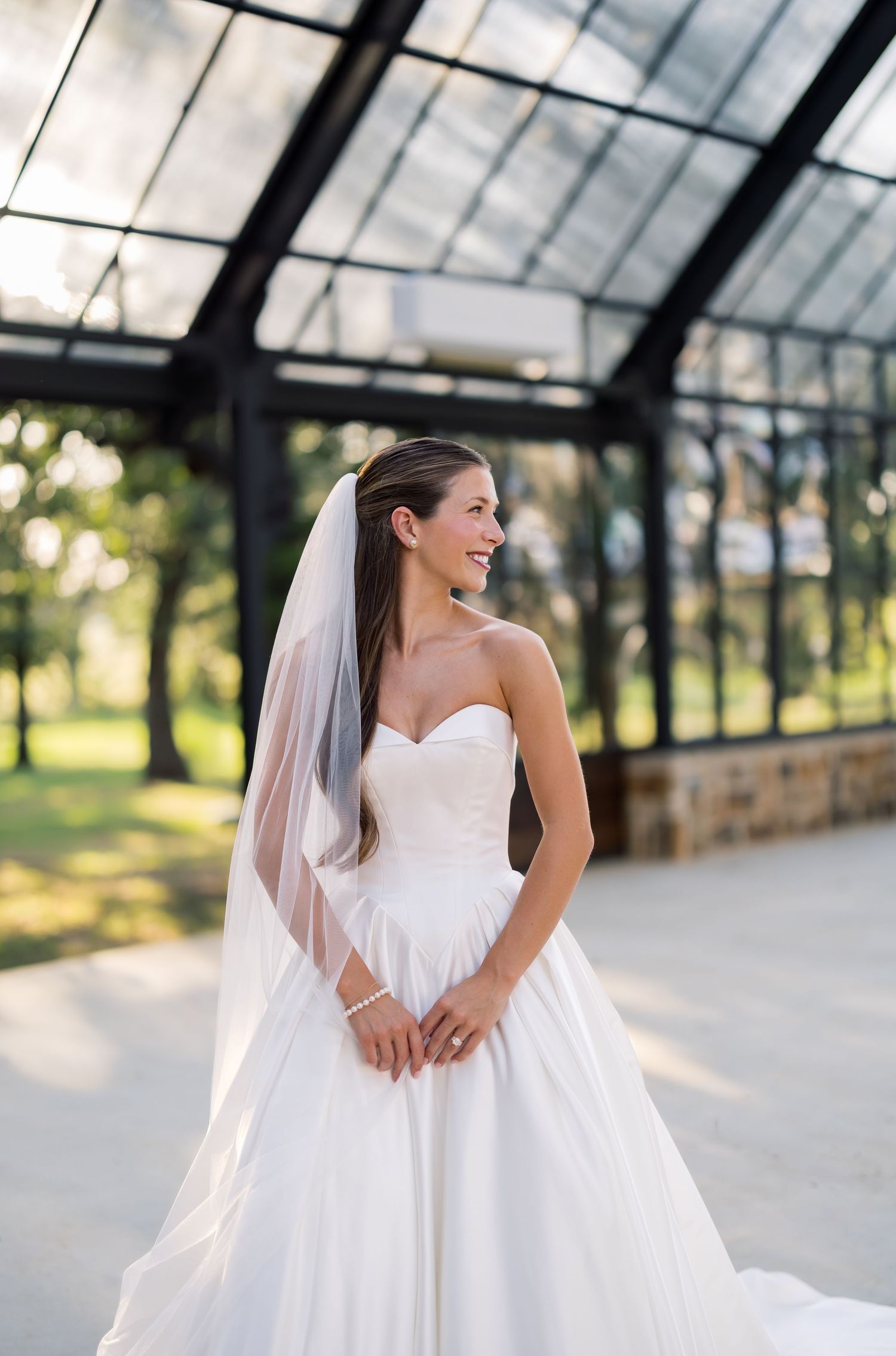 Smiling bride in strapless sweetheart ballgown with veil poses outdoors near glass pavilion.