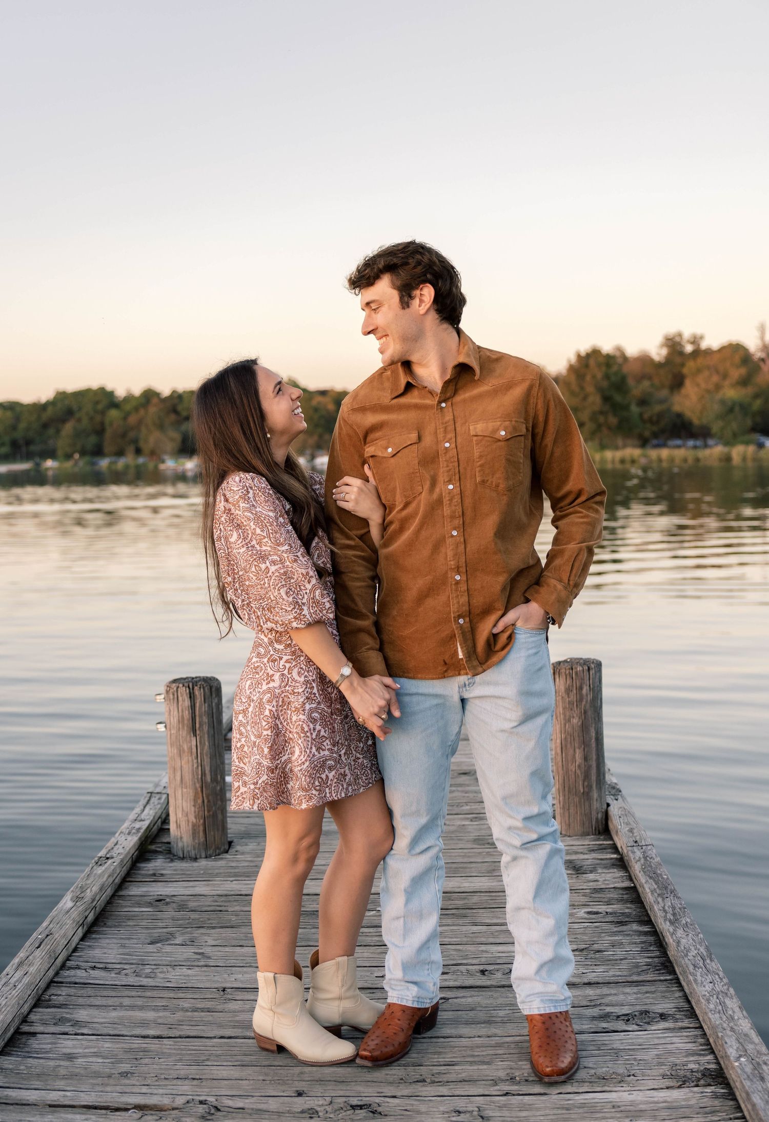 Couple laughing together on a lakeside dock at sunset, woman in floral dress and man in brown shirt.