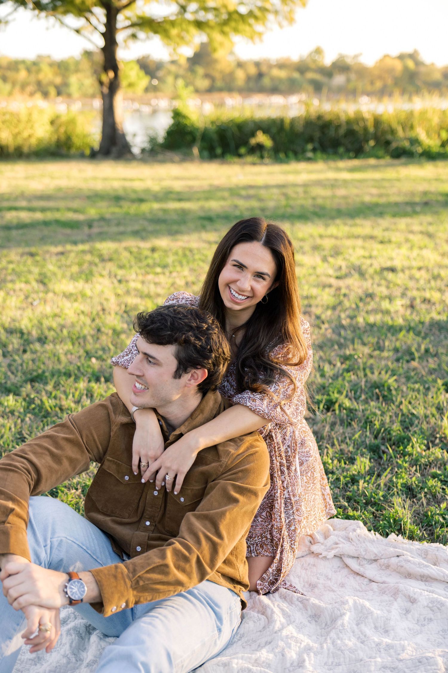 Woman embracing man from behind on a picnic blanket in a sunny park, both smiling joyfully.