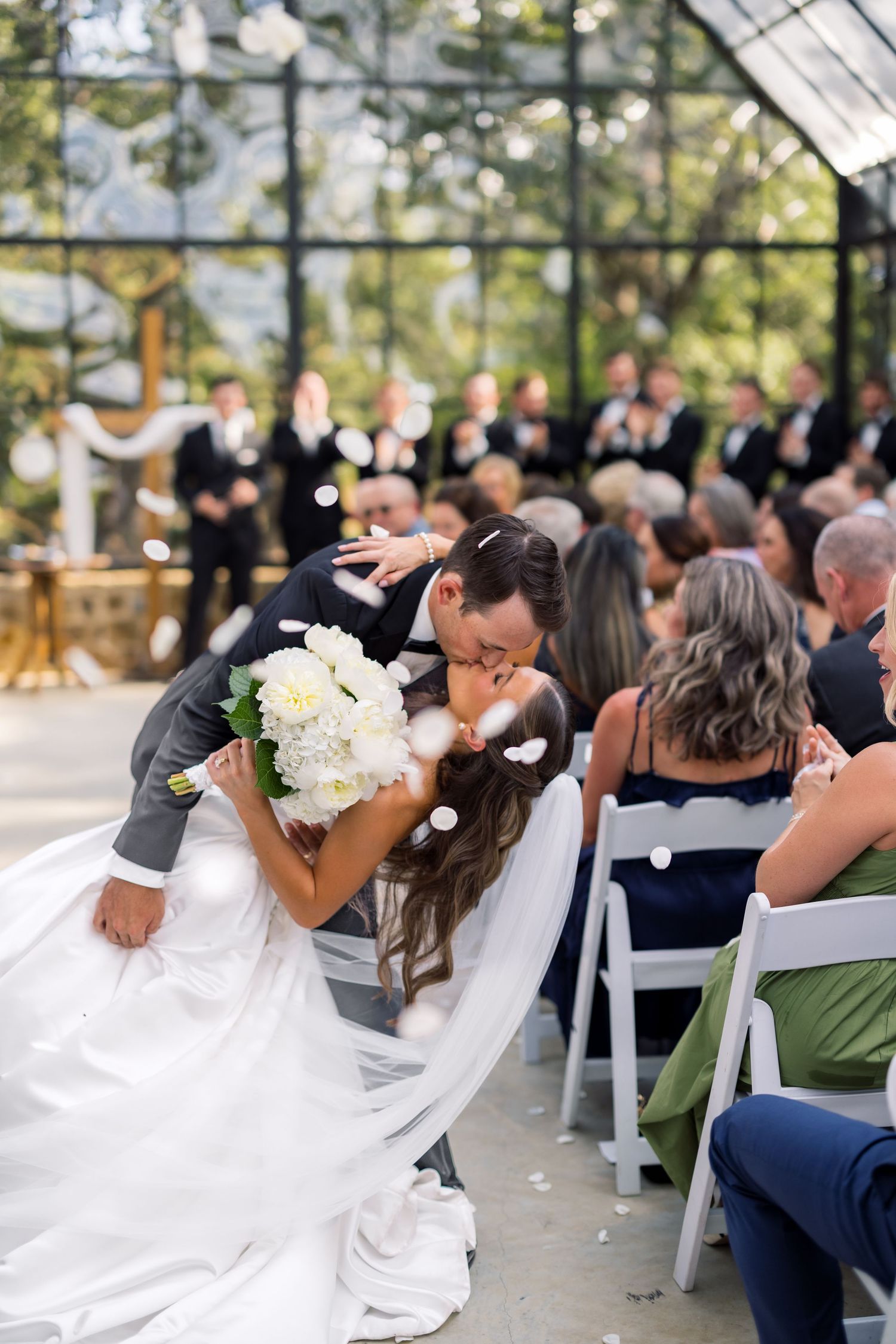 Bride and groom share a romantic dip kiss at wedding ceremony in greenhouse venue with guests seated nearby.