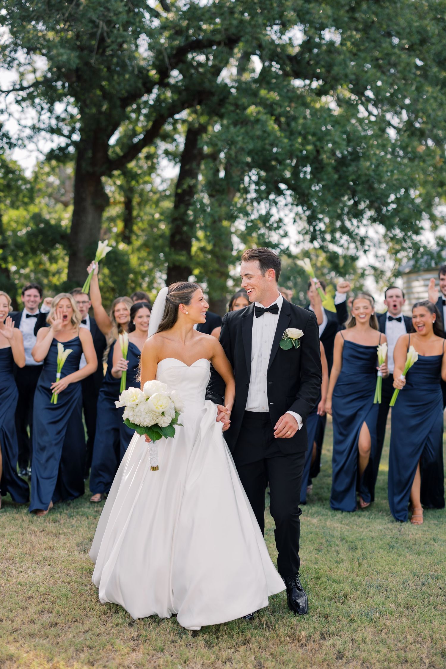 Bride in white gown and groom in black tuxedo walk together with wedding party in navy attire outdoors.