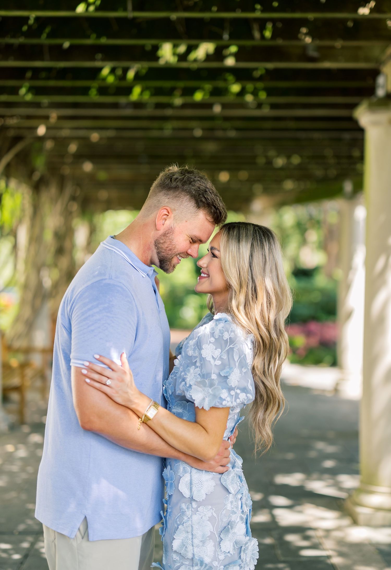 Couple embracing and smiling under a garden pergola, wearing coordinating blue outfits surrounded by lush greenery.