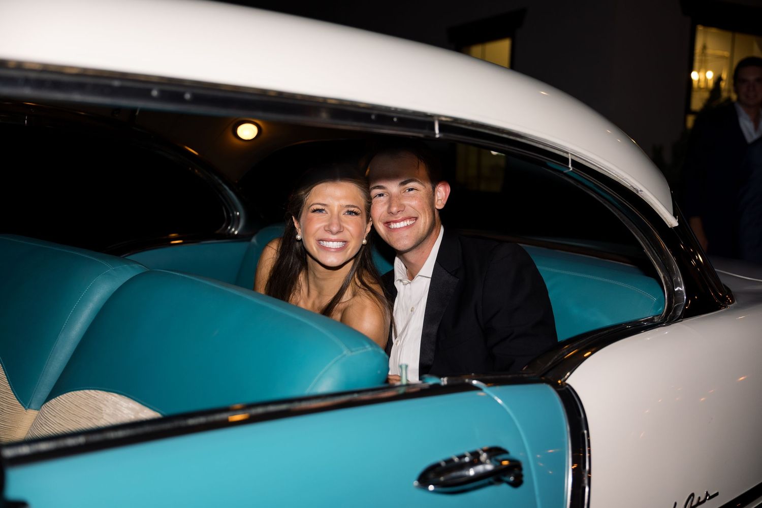 Smiling couple sitting in a classic teal and white vintage car interior at a formal evening event.