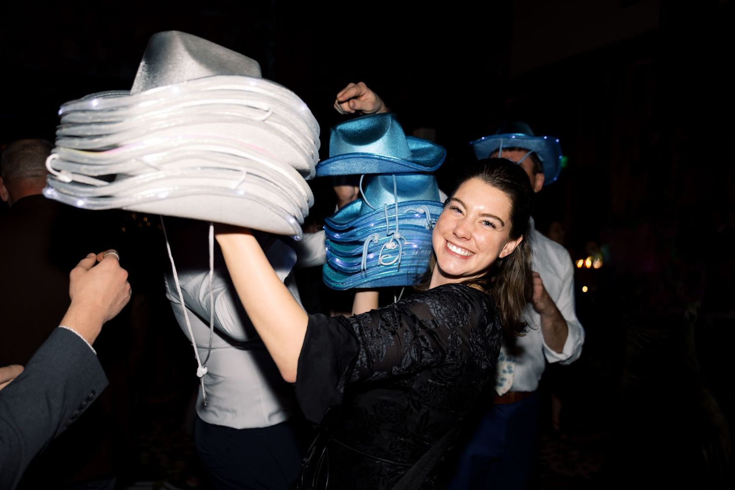 A smiling woman enjoys a lively party, holding blue and white items above her head in a festive indoor setting.