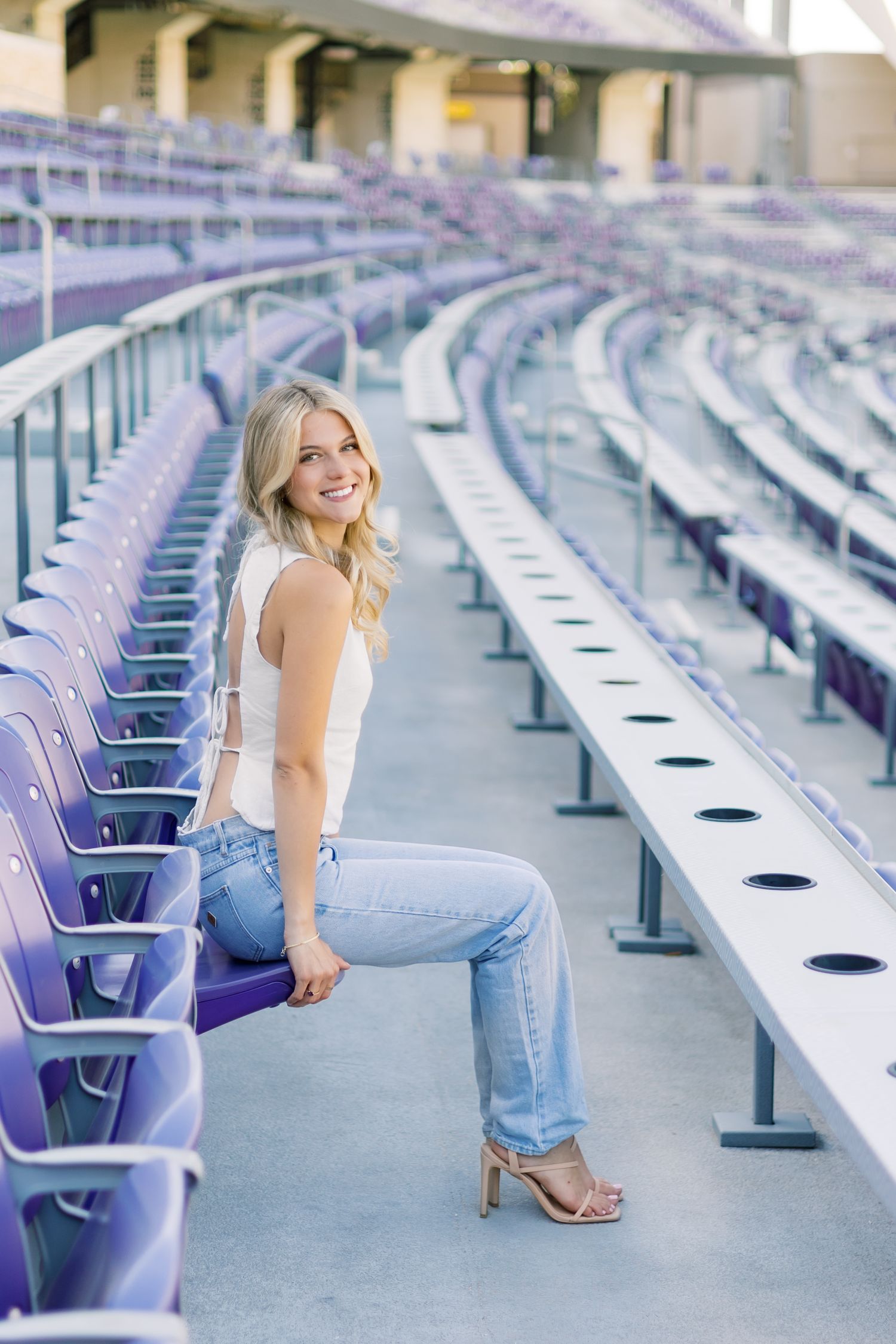 Smiling blonde woman in jeans and white top sitting in empty purple stadium seats.