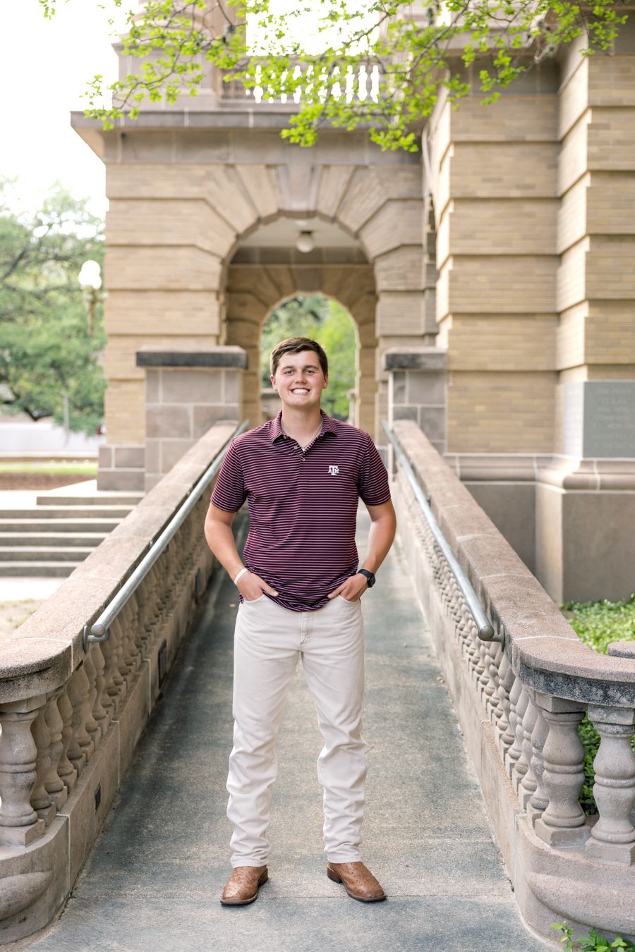 Young man in maroon polo and khakis smiling on a stone bridge walkway outside a classic university building.