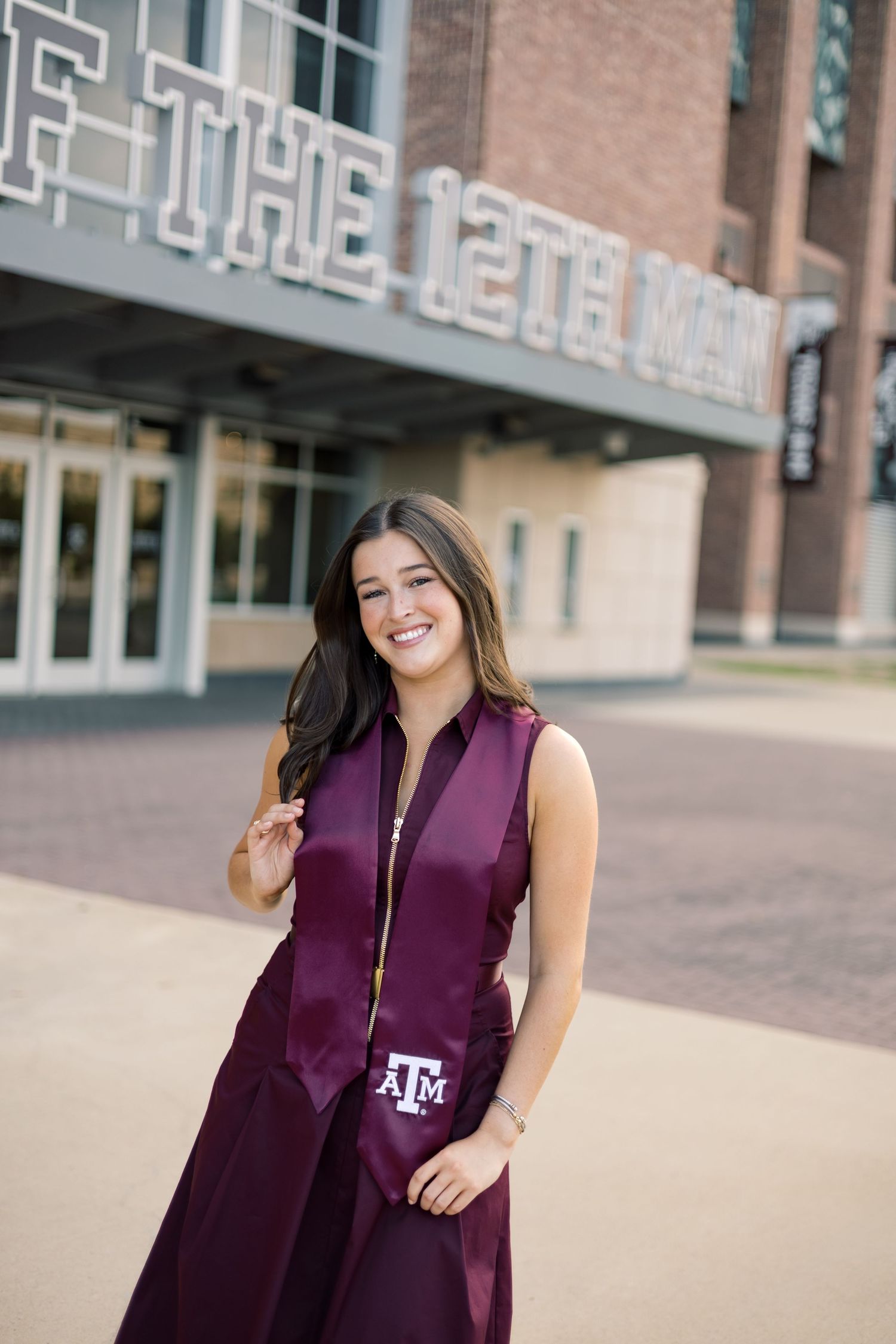 Texas A&M graduate smiling in maroon cap and gown outside Kyle Field, home of the 12th Man.