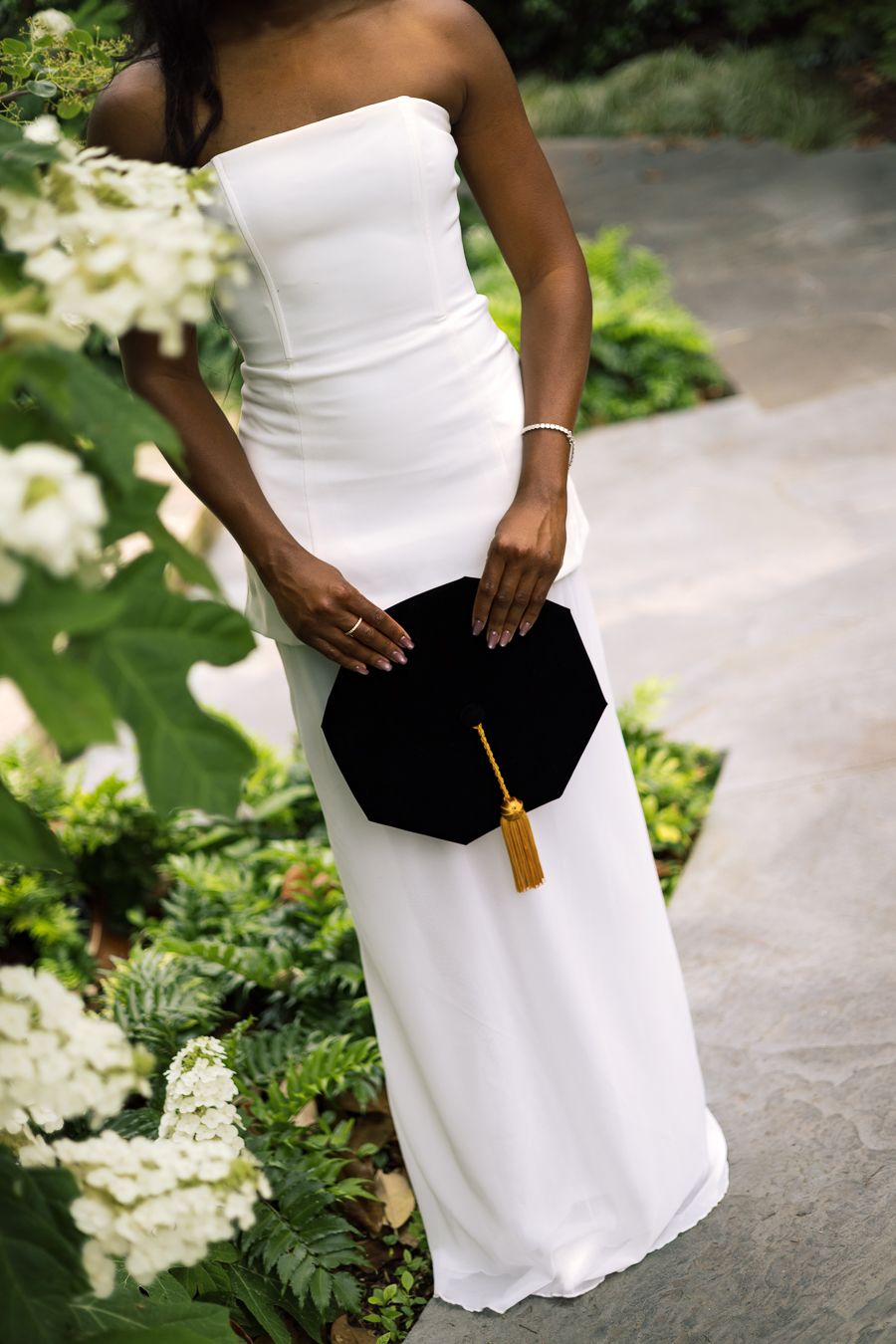 Black woman in white strapless gown holding graduation cap with gold tassel among white flowers.