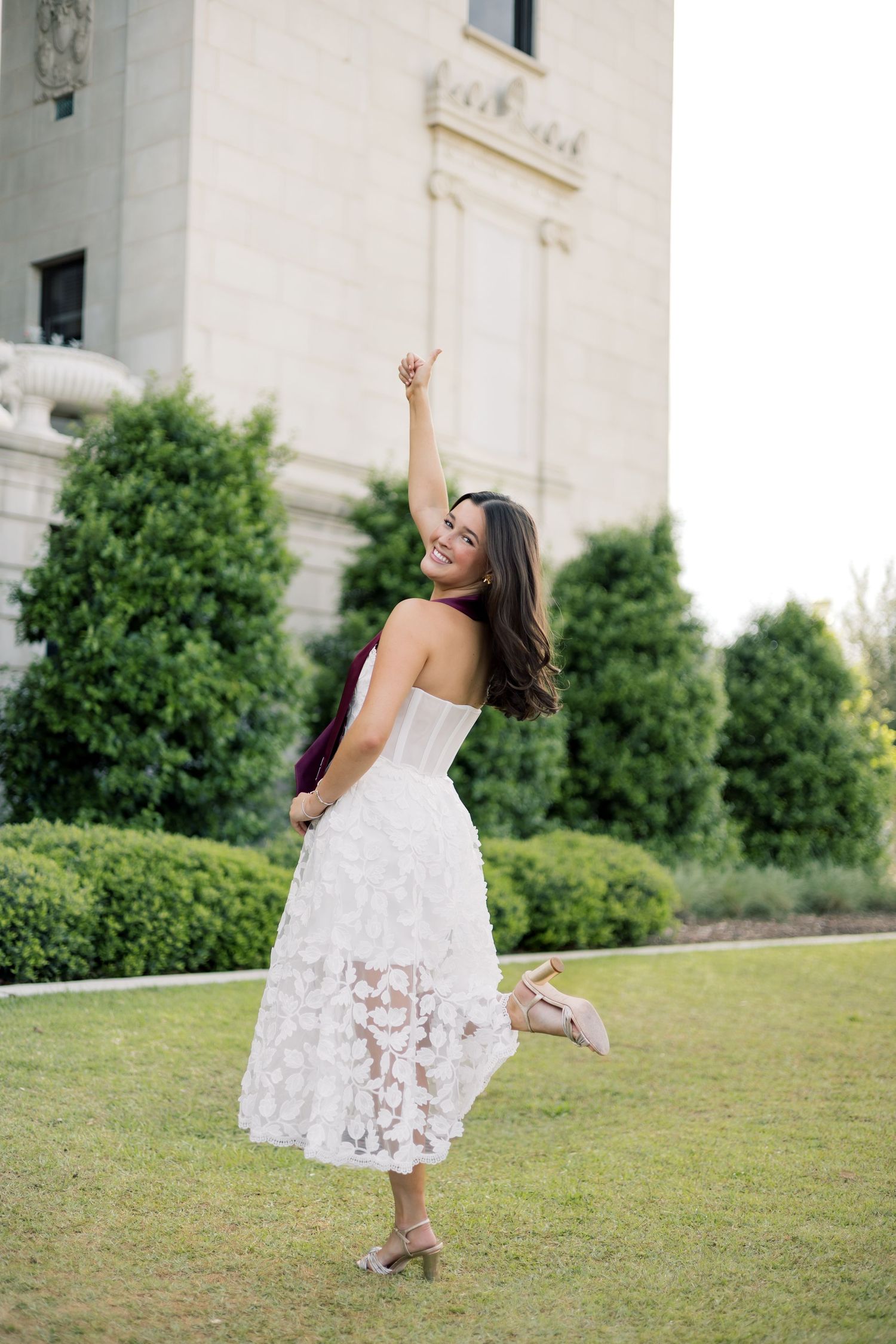 Joyful graduate in white lace dress raising hand and holding heel while spinning on campus lawn.