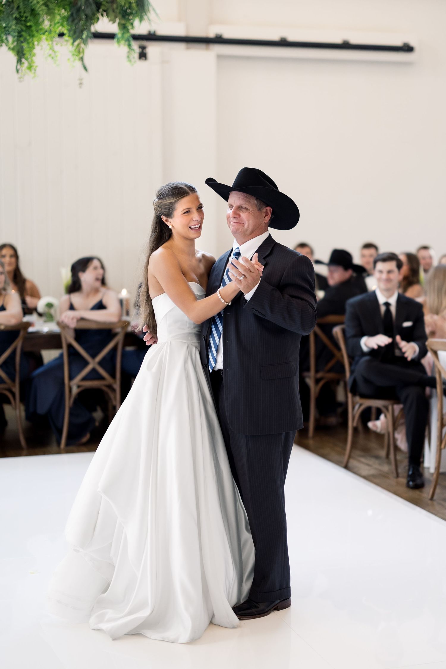 Bride in white gown dances with man in black suit and cowboy hat at wedding reception on white dance floor.