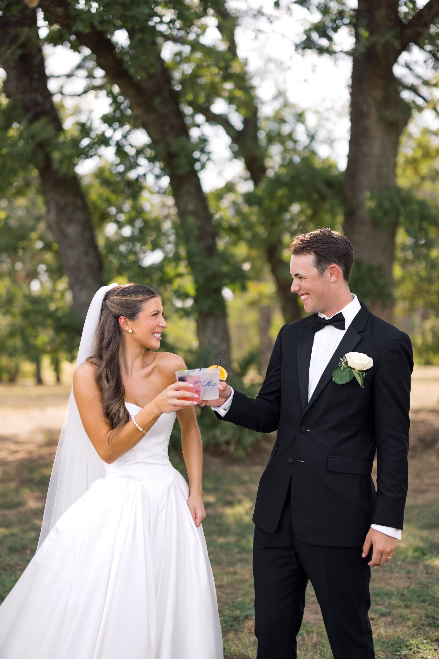 Bride and groom toasting with cocktails outdoors under oak trees, wearing wedding attire with white rose boutonniere.