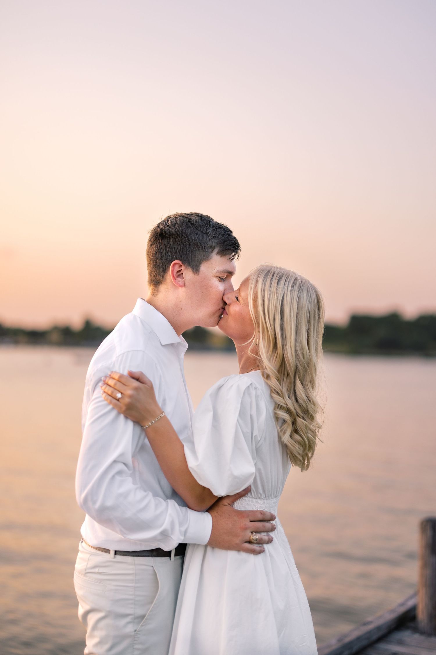 Couple sharing a romantic kiss at sunset by a lake, dressed in white outfits on a wooden dock.