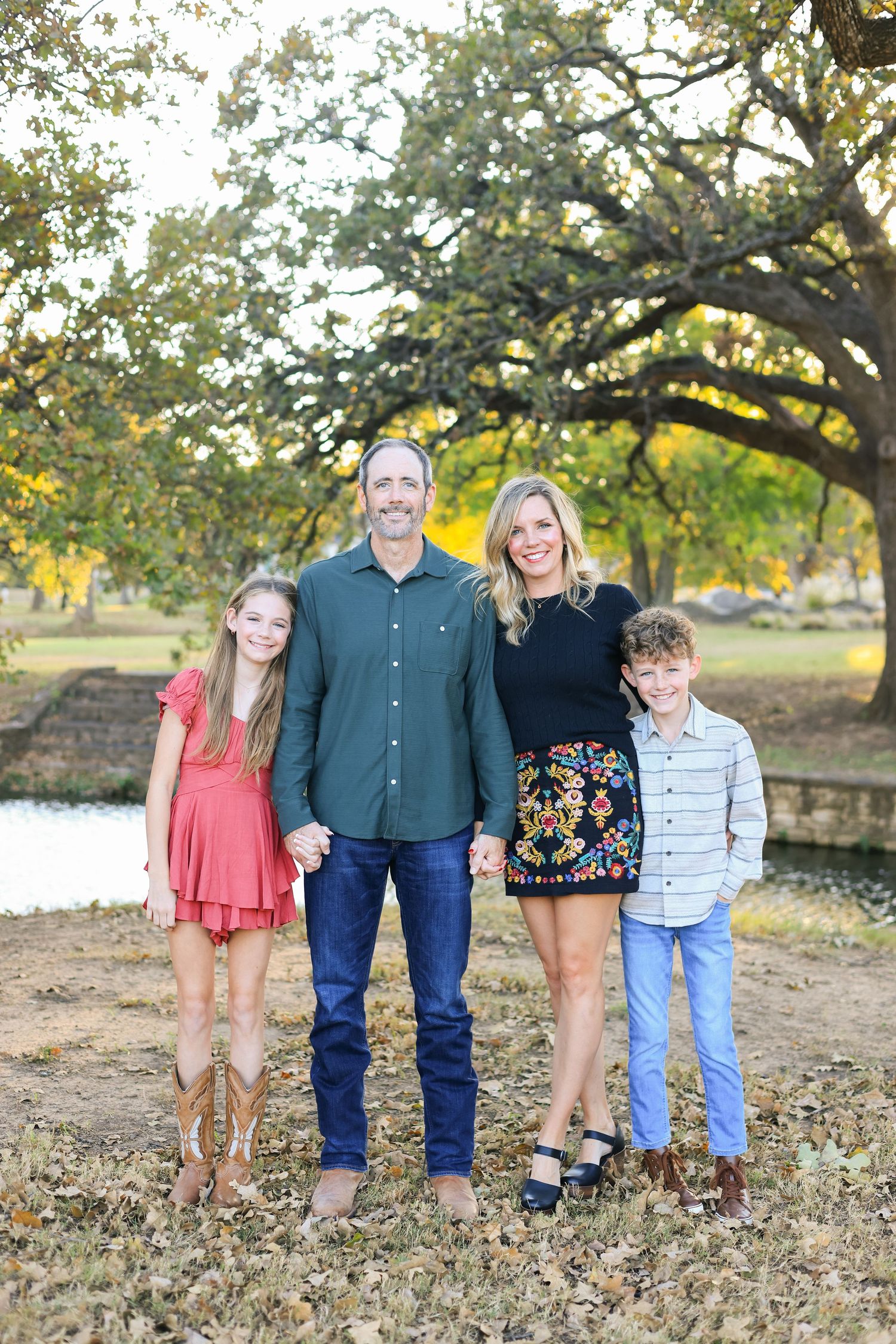Family of four smiling outdoors under large oak trees, parents with daughter in red dress and son in plaid shirt.