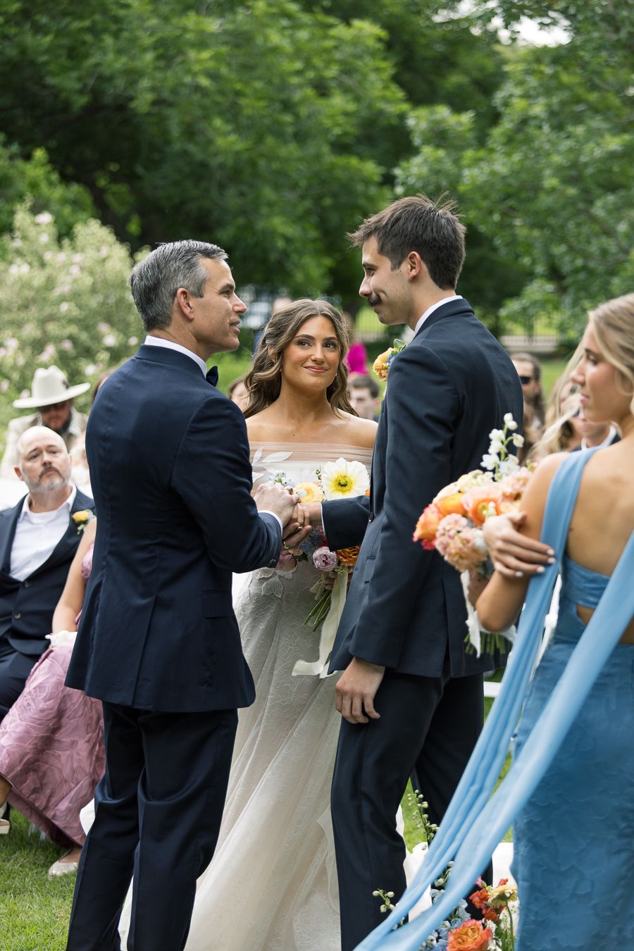 Outdoor wedding ceremony with bride in white gown, groom in navy suit shaking hands with man, guests seated on green lawn.
