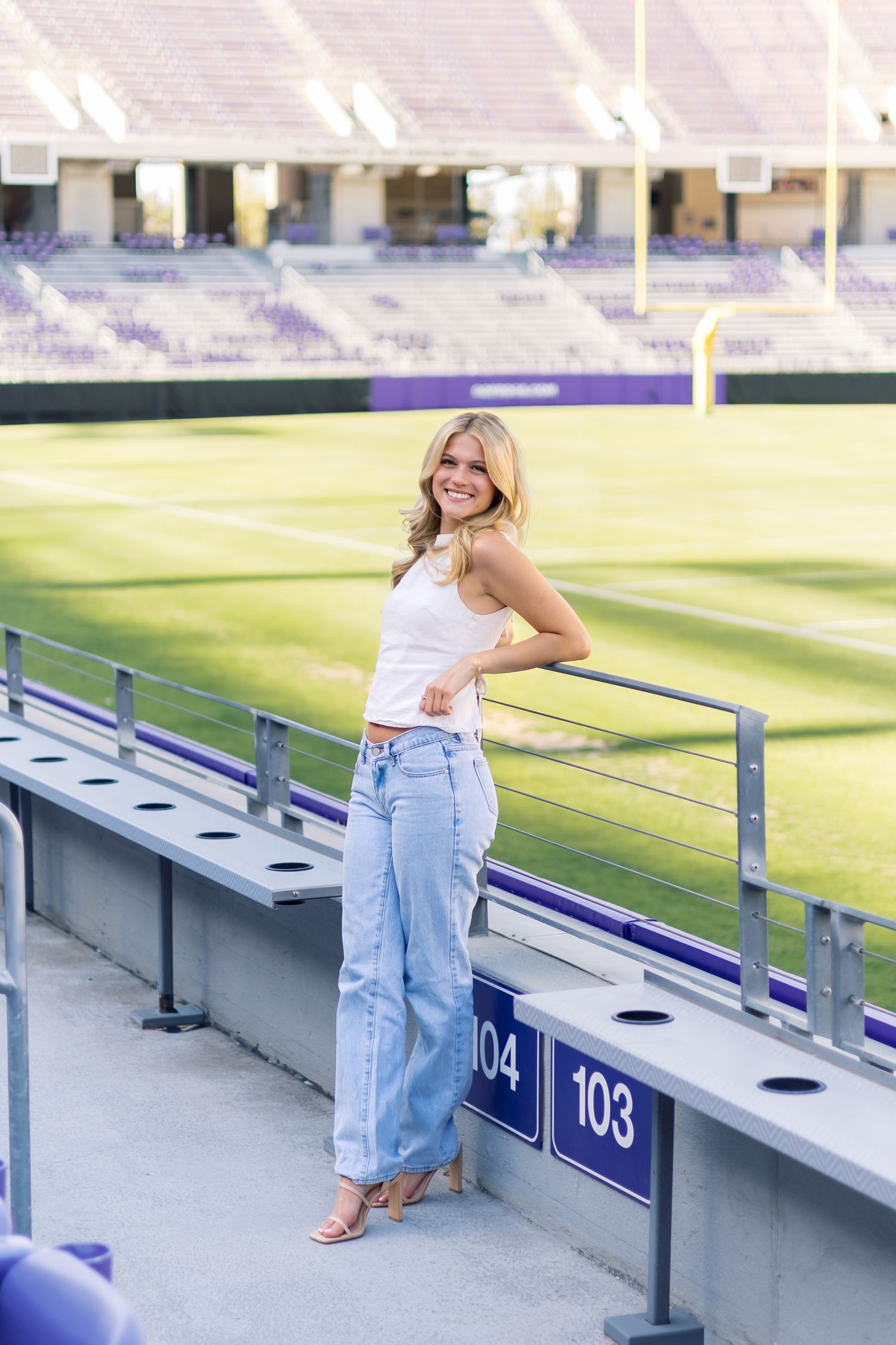 Smiling blonde woman in white top and wide-leg jeans leans on railing at sections 103-104 in stadium.