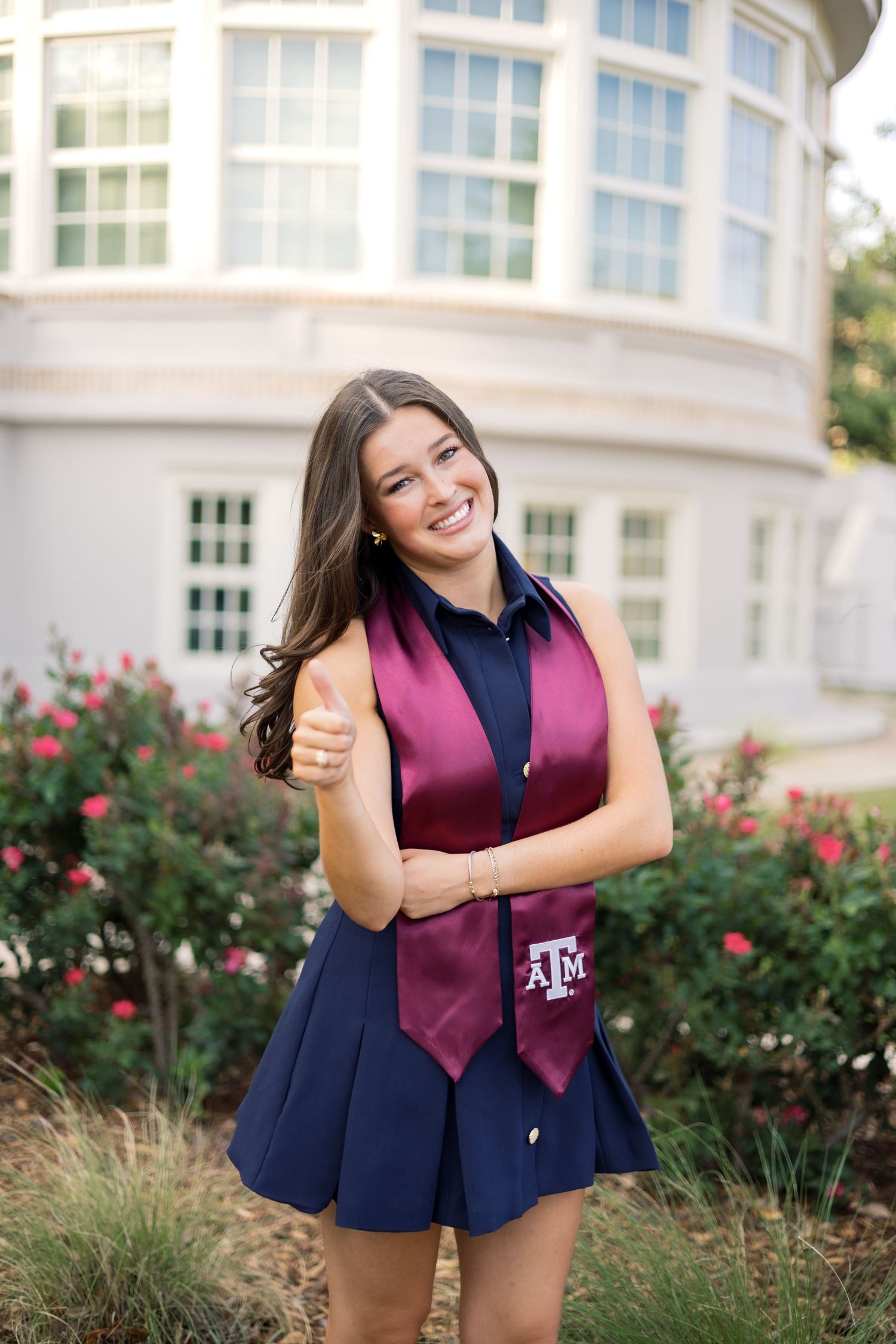 Cheerful brunette in navy dress and Texas A&M maroon stole giving thumbs up in front of a building with roses.