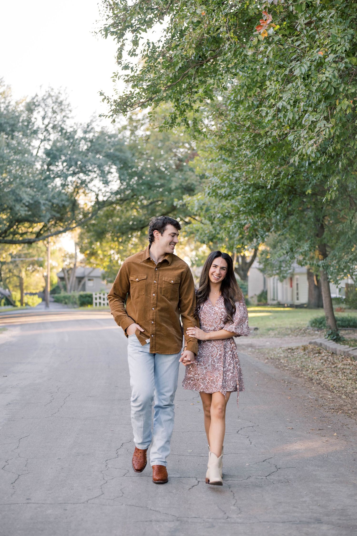 Couple holding hands walking down a tree-lined path, man in brown jacket and woman in floral dress with white boots.