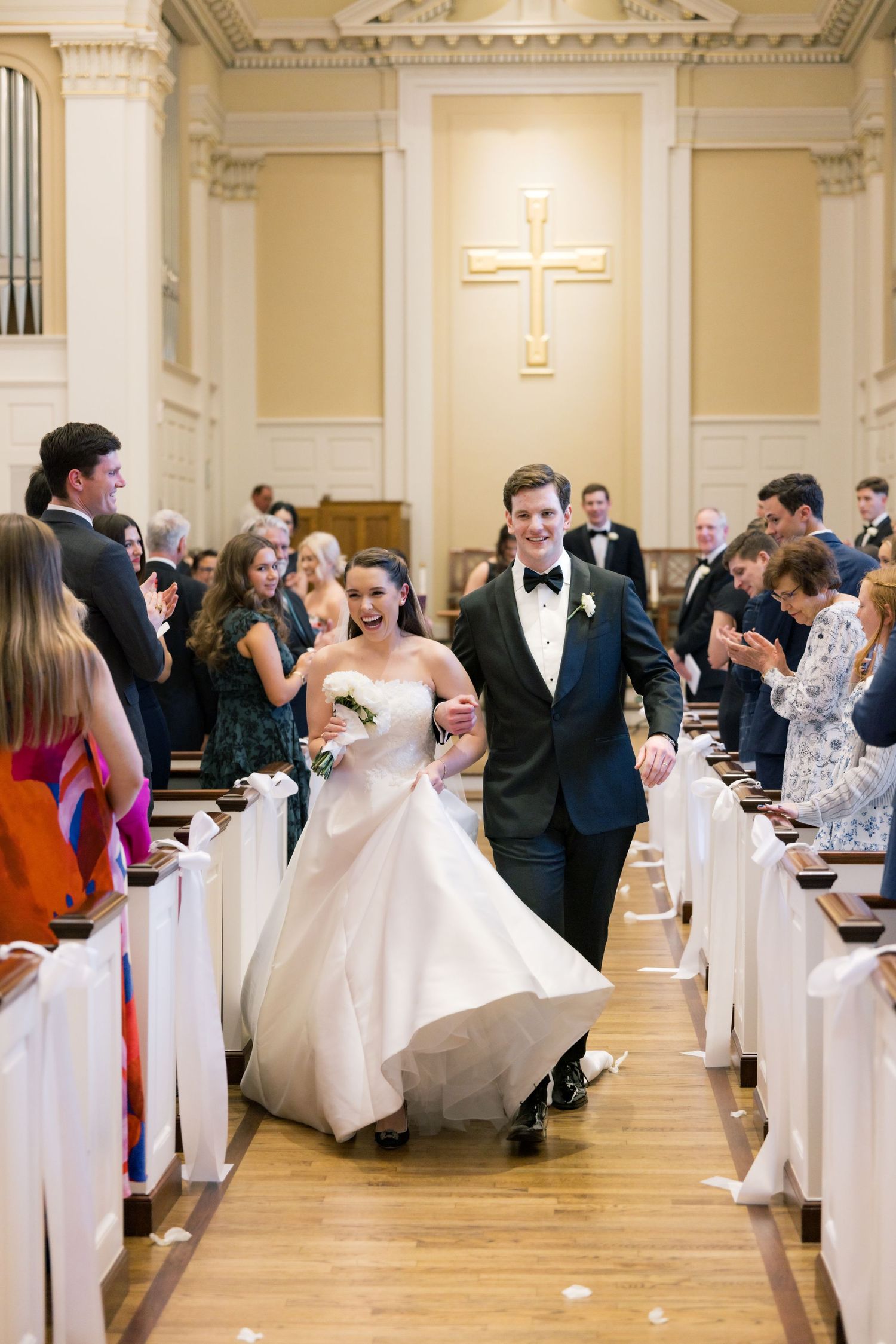 Bride and groom walk down church aisle after wedding ceremony, guests applauding, cross visible in background.