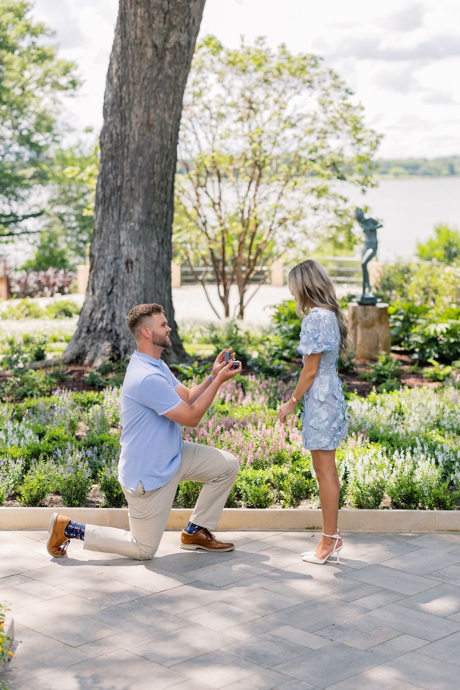 Man proposing on one knee to woman in floral dress in a scenic garden with a bronze statue and waterfront.