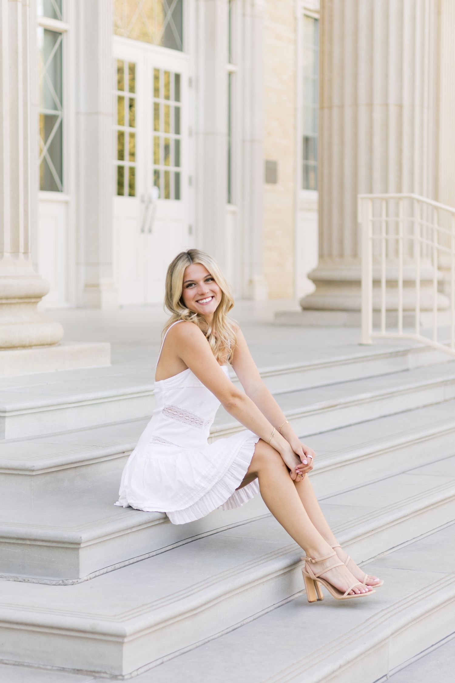Smiling blonde woman in white sundress sitting on marble steps of neoclassical university building.