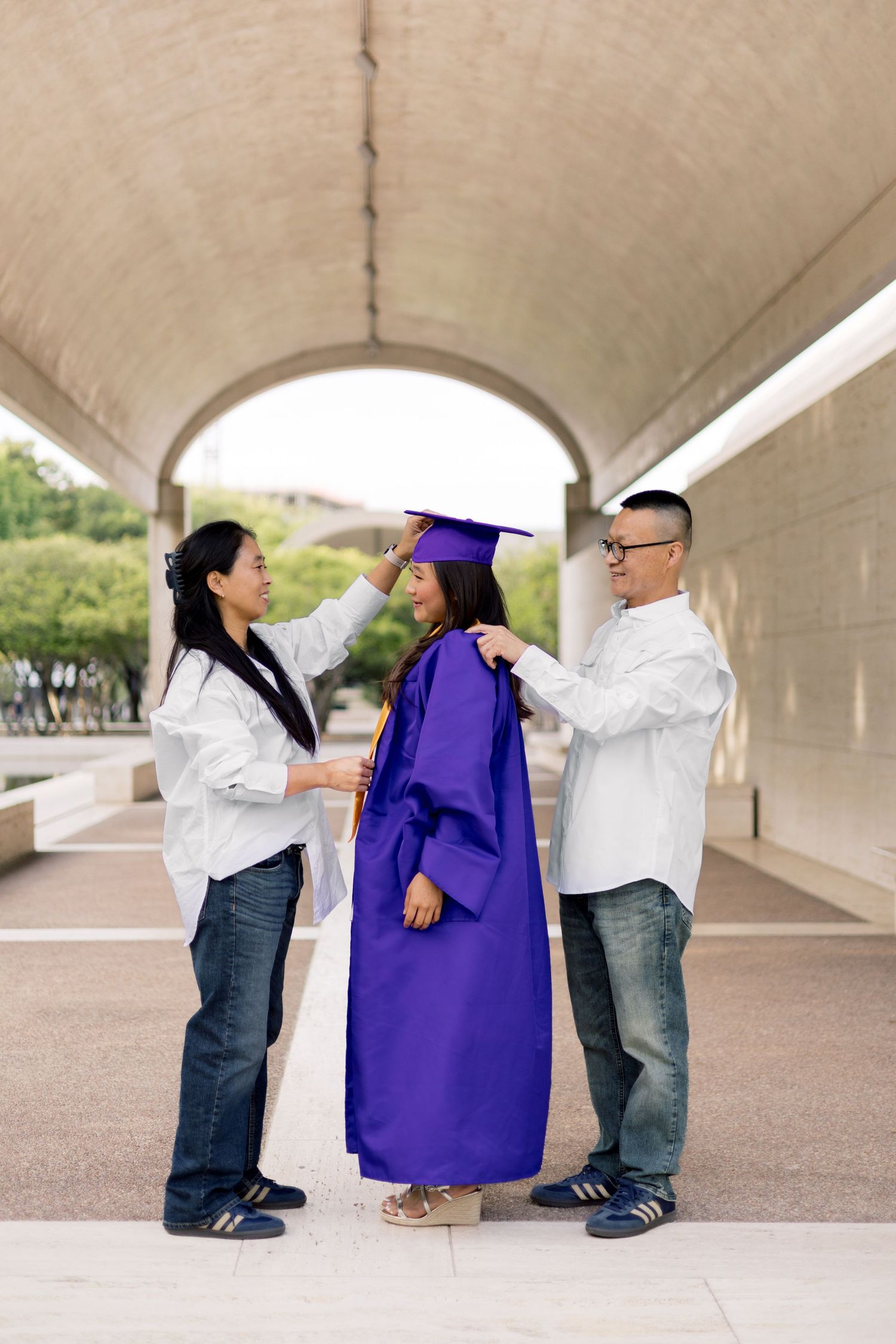 Parents helping their daughter in purple graduation cap and gown prepare for her ceremony outdoors.