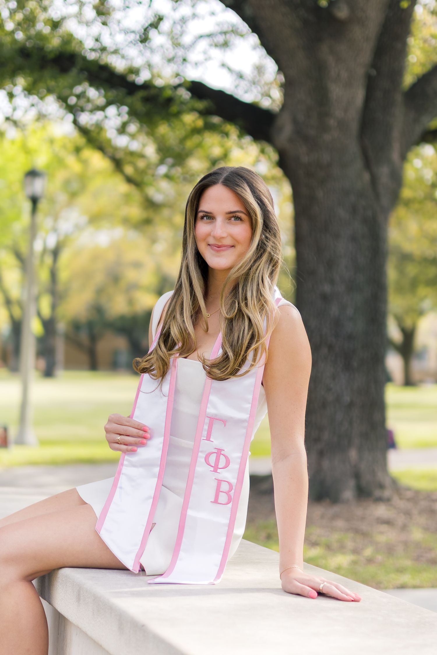 Young woman wearing a Gamma Phi Beta sorority sash poses outdoors on a stone ledge near a large tree.