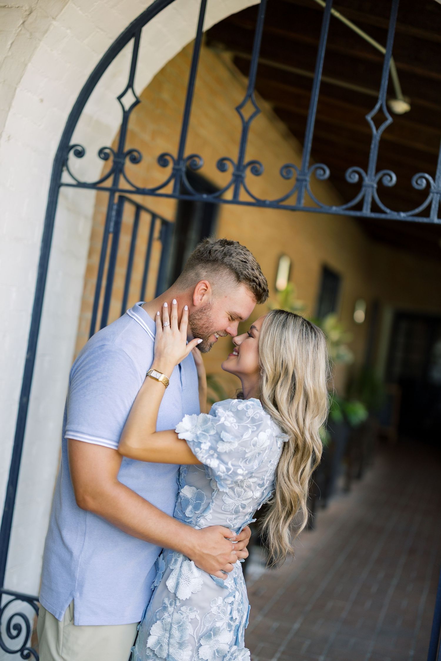 Couple sharing a romantic moment under an ornate iron archway, woman in floral dress, man in blue polo shirt.