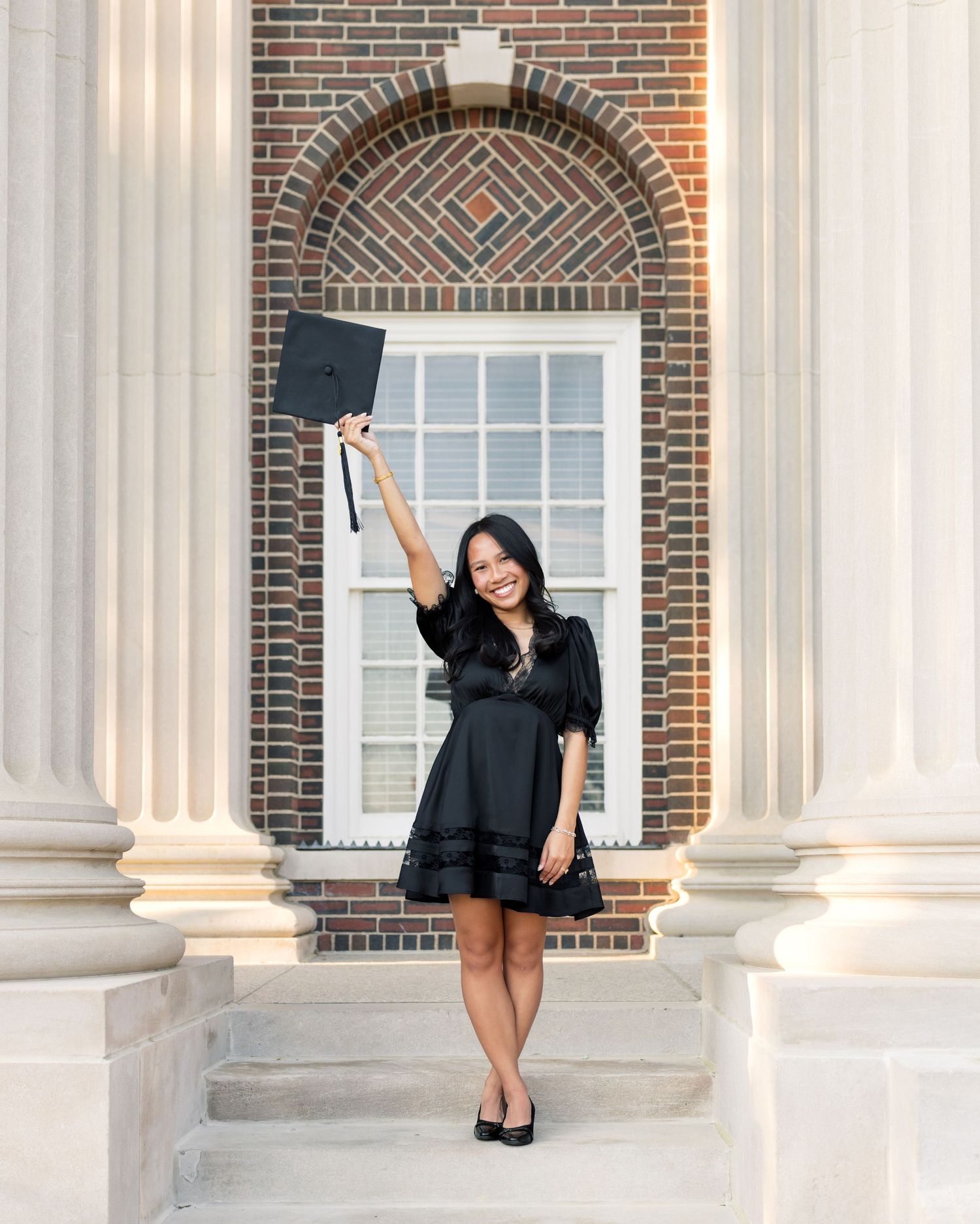 Young woman in black dress celebrating graduation by tossing cap in front of brick building with columns.