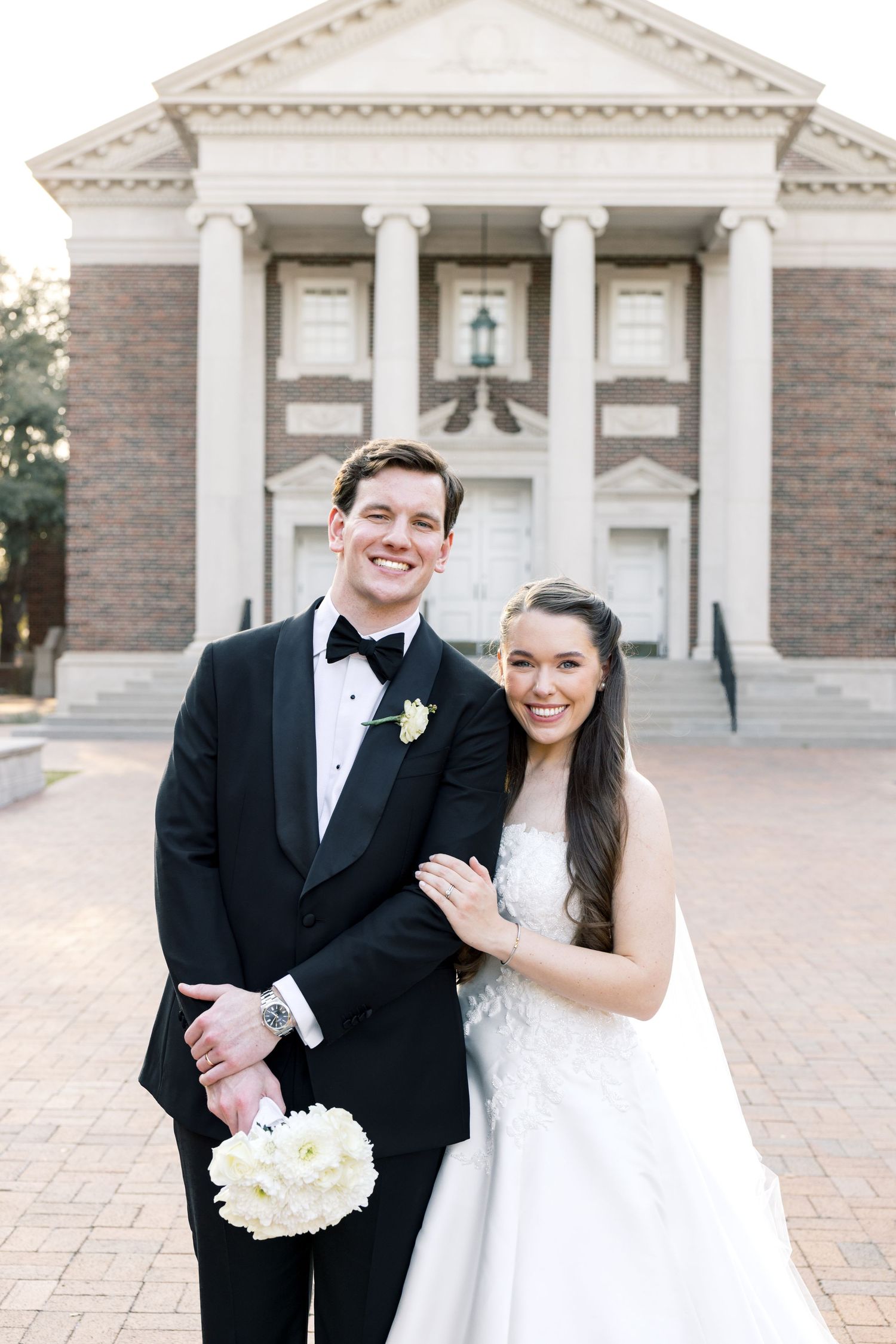 Smiling groom in black tuxedo and bride in white gown pose together before a classic brick building.