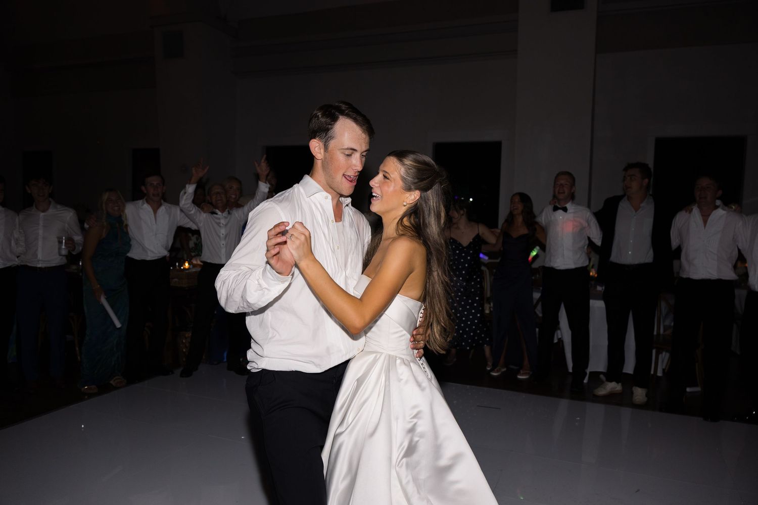Bride and groom share their first dance at wedding reception with guests watching in background.