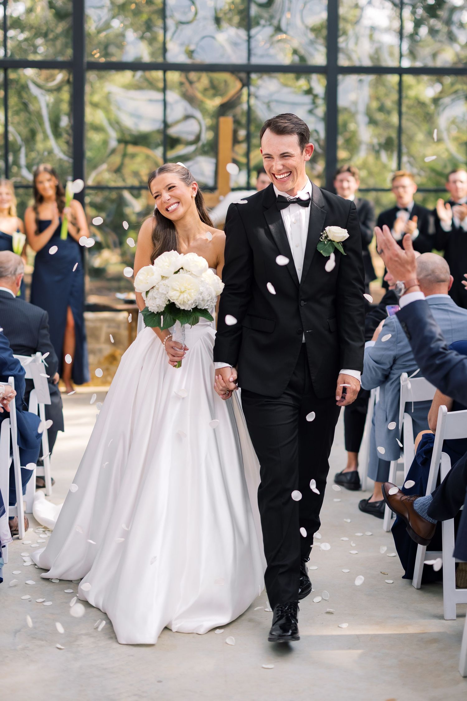 Bride and groom walk down the aisle smiling, showered with white petals, bride holding white bouquet.