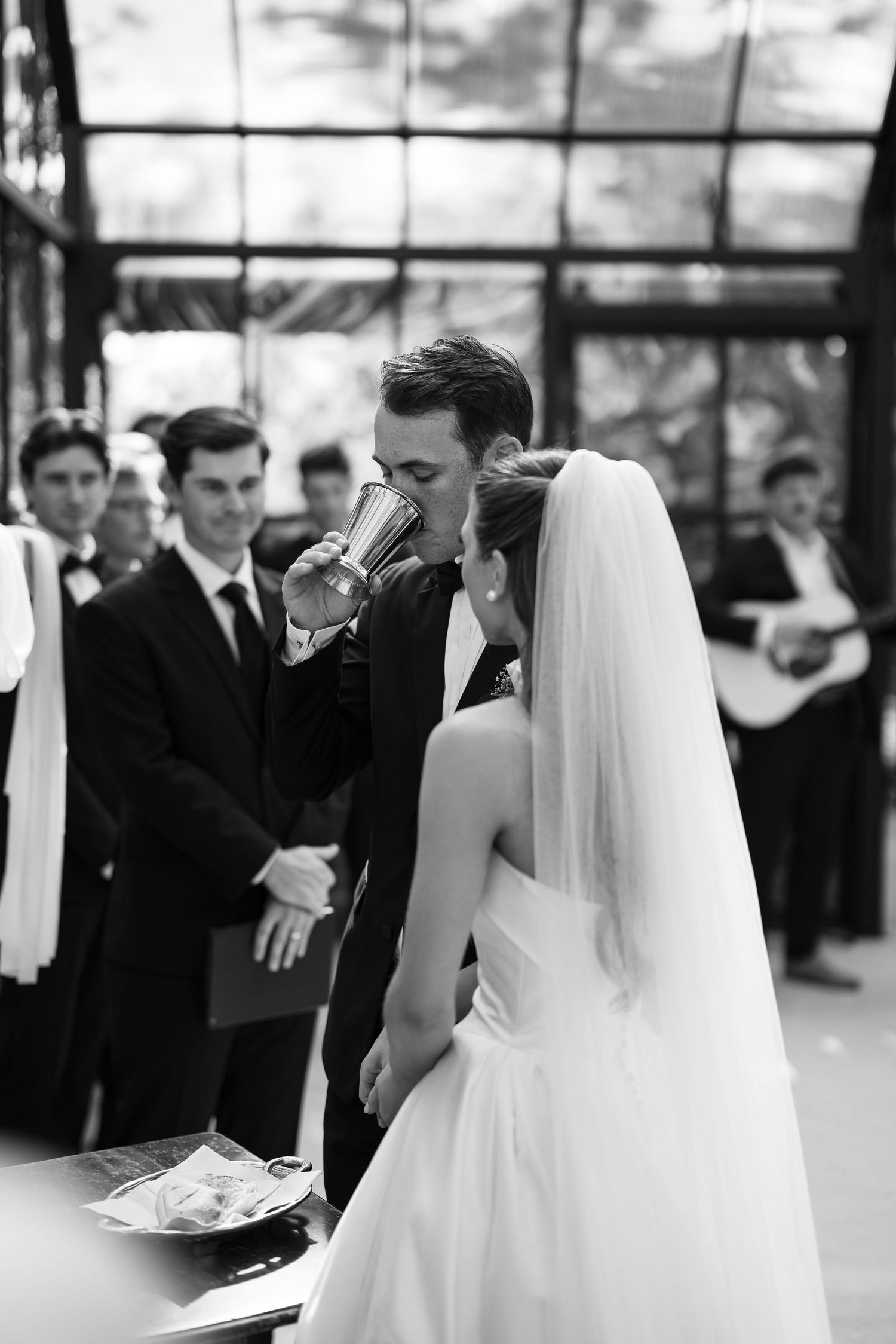 Black and white wedding photo of bride and groom sharing a ceremonial cup drink at their elegant indoor wedding ceremony.
