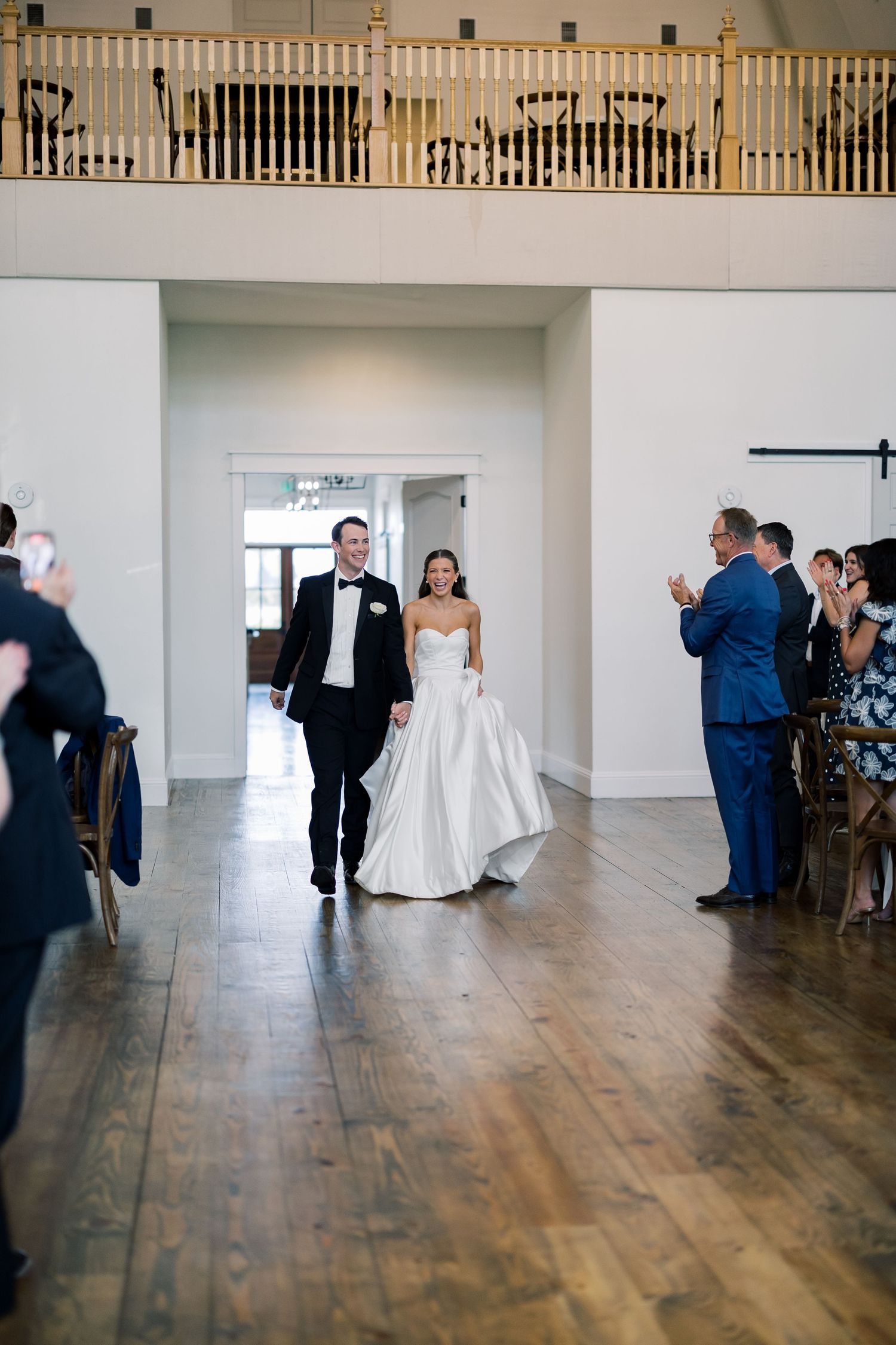 Bride and groom walk down the aisle at their elegant wedding reception while guests applaud in a bright venue.