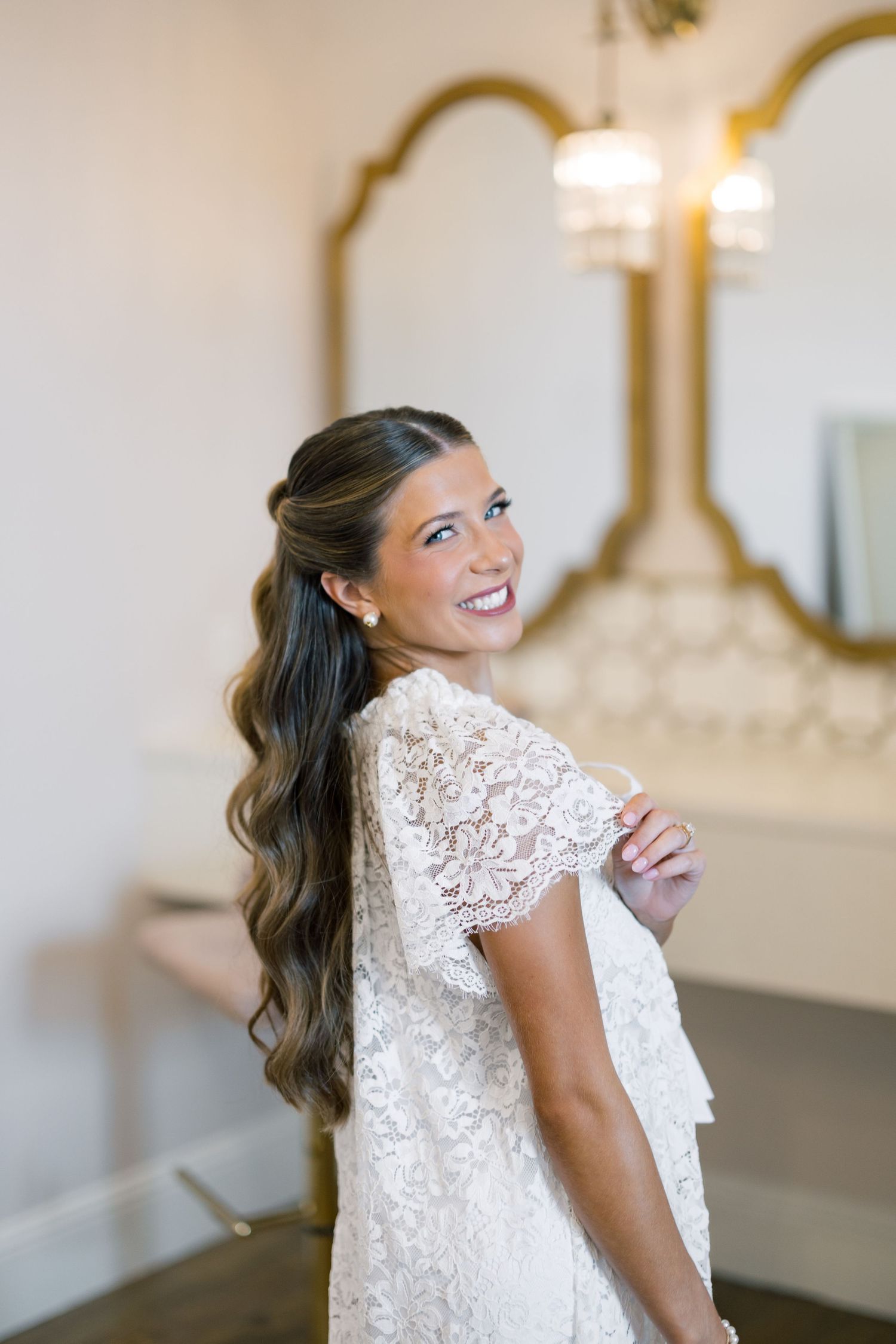Smiling bride in lace wedding dress with half-up wavy hairstyle posing in elegant bridal suite with gold mirror.