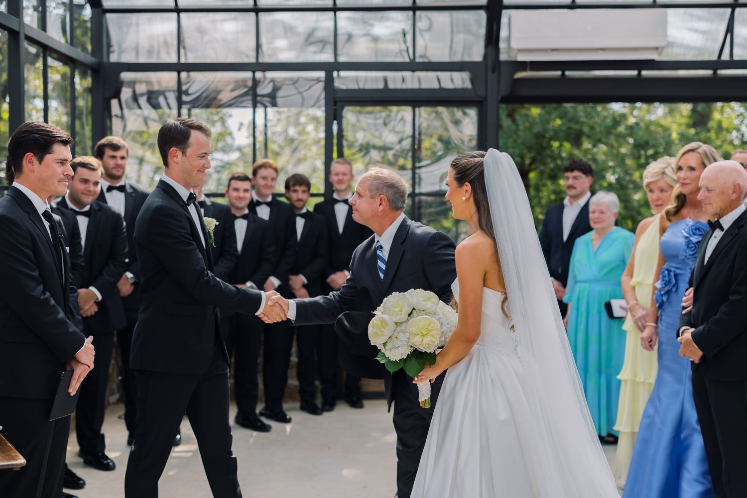 Groom shaking hands with father of the bride at elegant indoor wedding ceremony with wedding party.