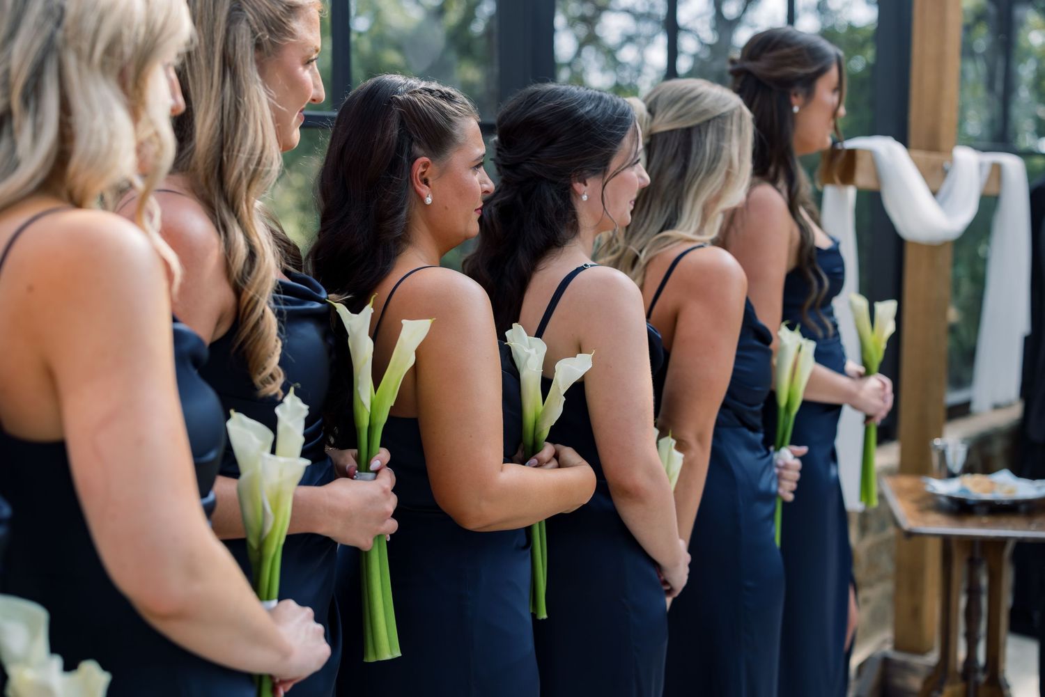 Bridesmaids in navy blue dresses holding white calla lily bouquets stand in a row during a wedding ceremony.