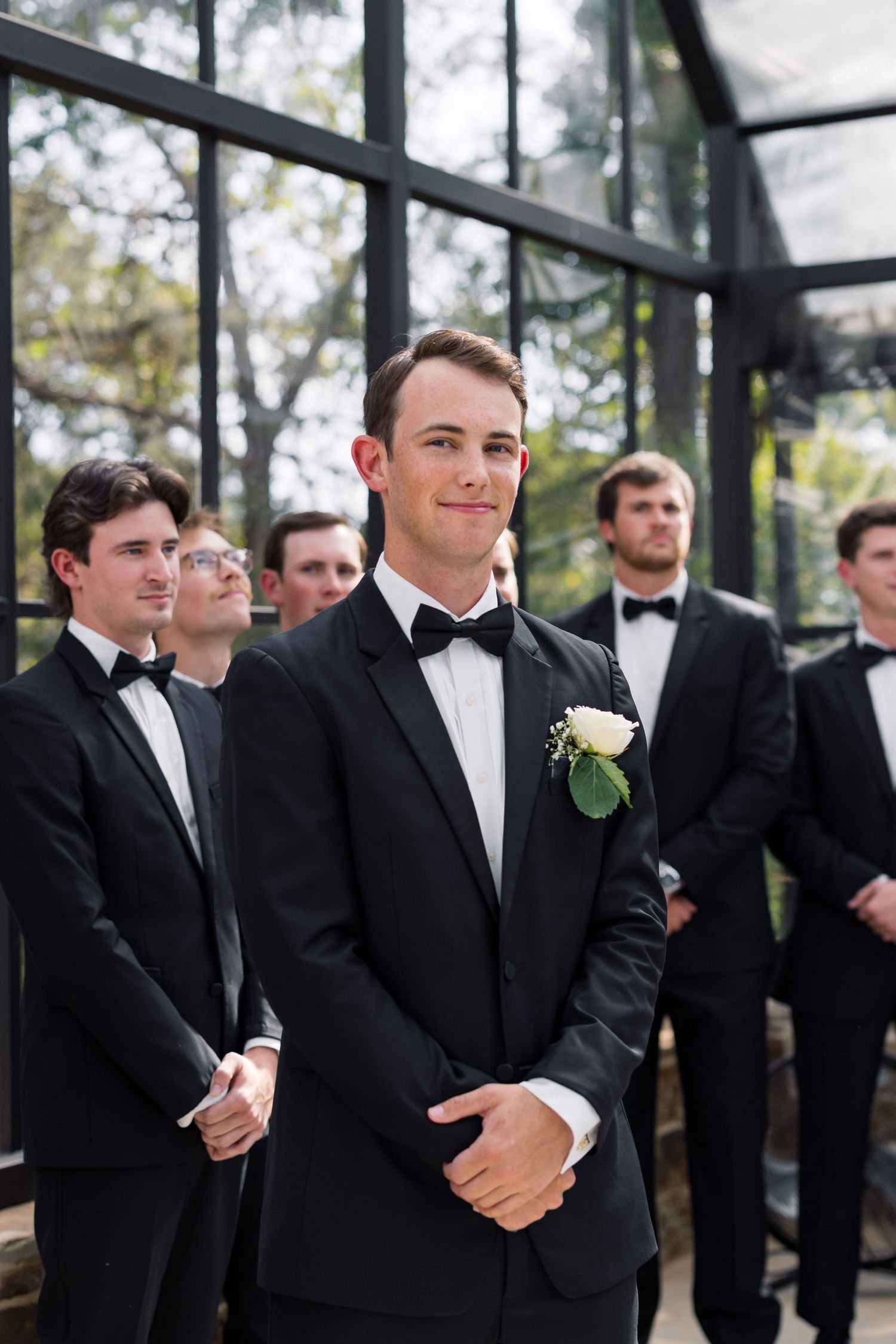 Groom in black tuxedo with white boutonniere stands before groomsmen at outdoor wedding ceremony.