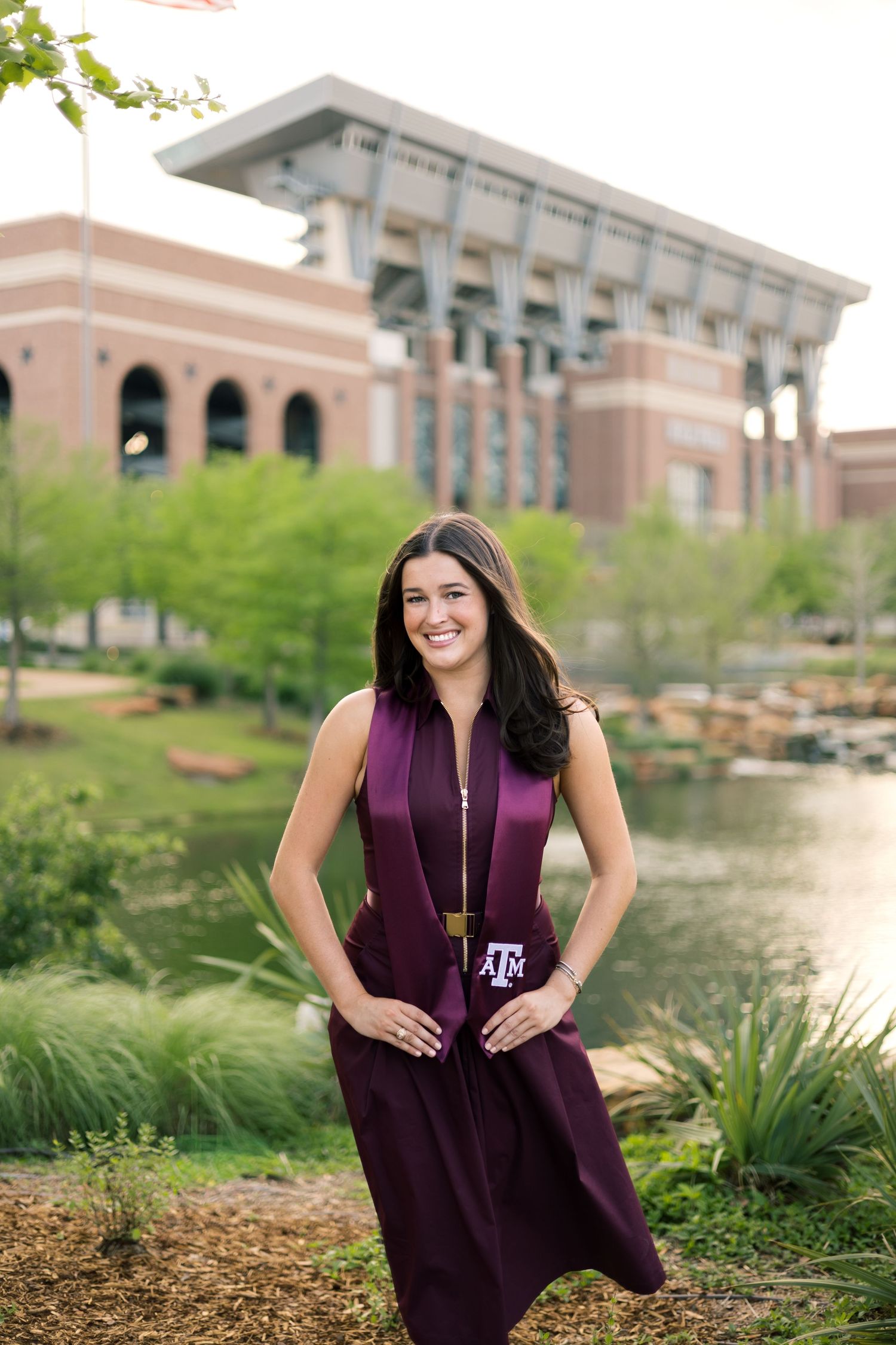 Smiling Texas A&M grad holds stole near scenic pond with stadium backdrop on campus.