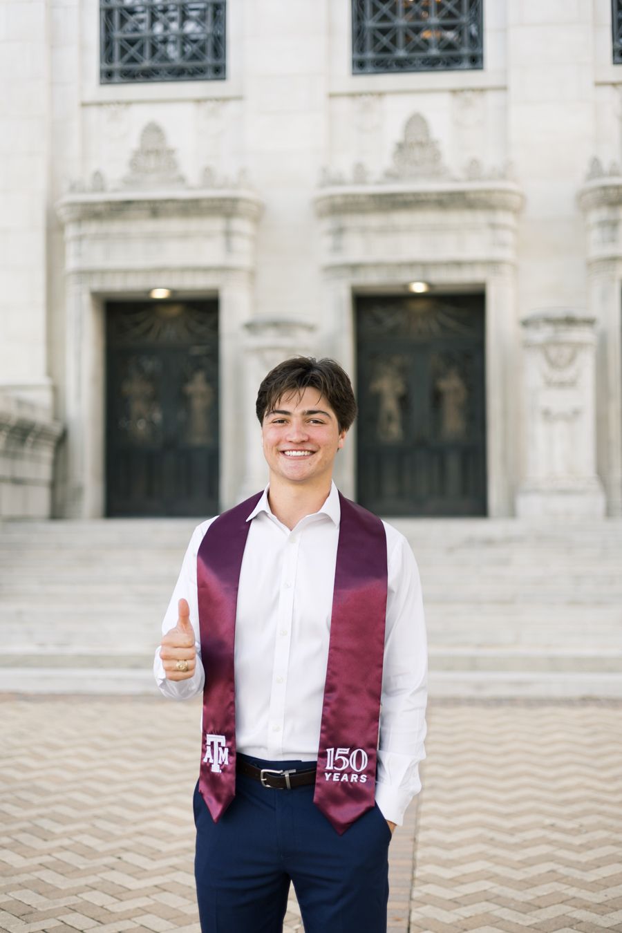 Young man in white shirt with maroon Texas A&M 150 Years graduation stole giving thumbs up outside university building.