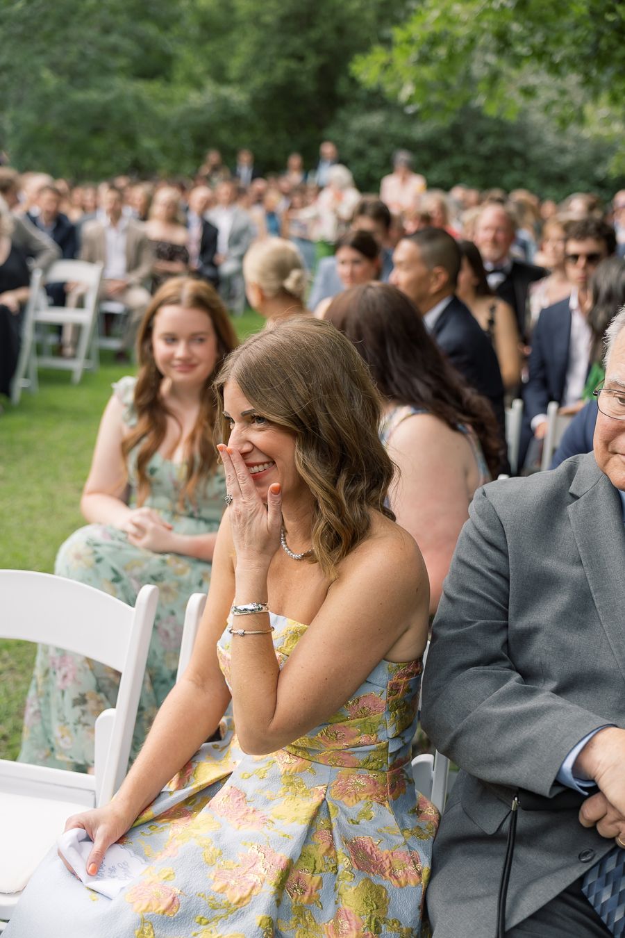 A smiling wedding guest in a floral dress laughs joyfully during an outdoor garden wedding ceremony.