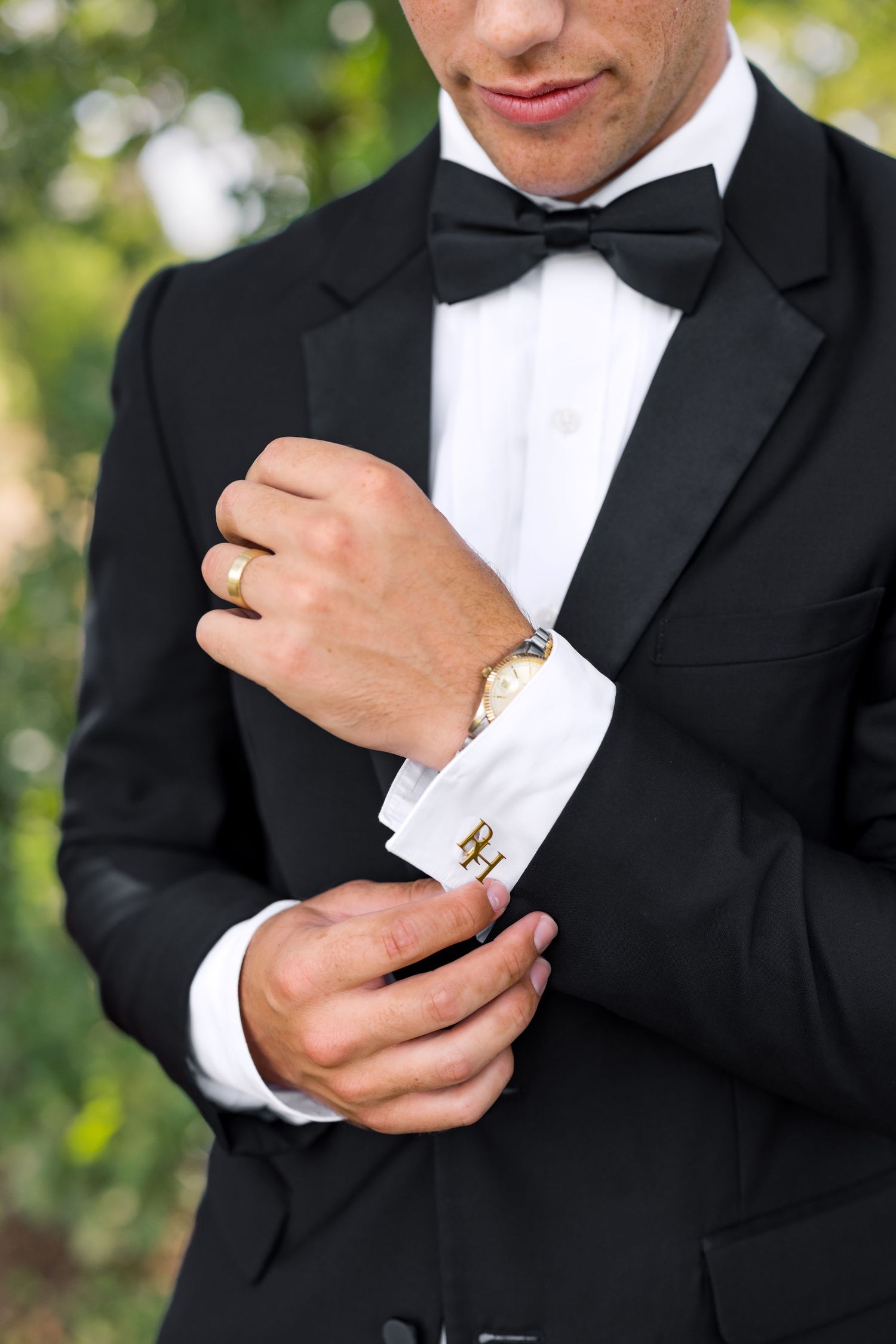Man in black tuxedo with bow tie adjusting gold cufflinks, wearing a watch and ring.