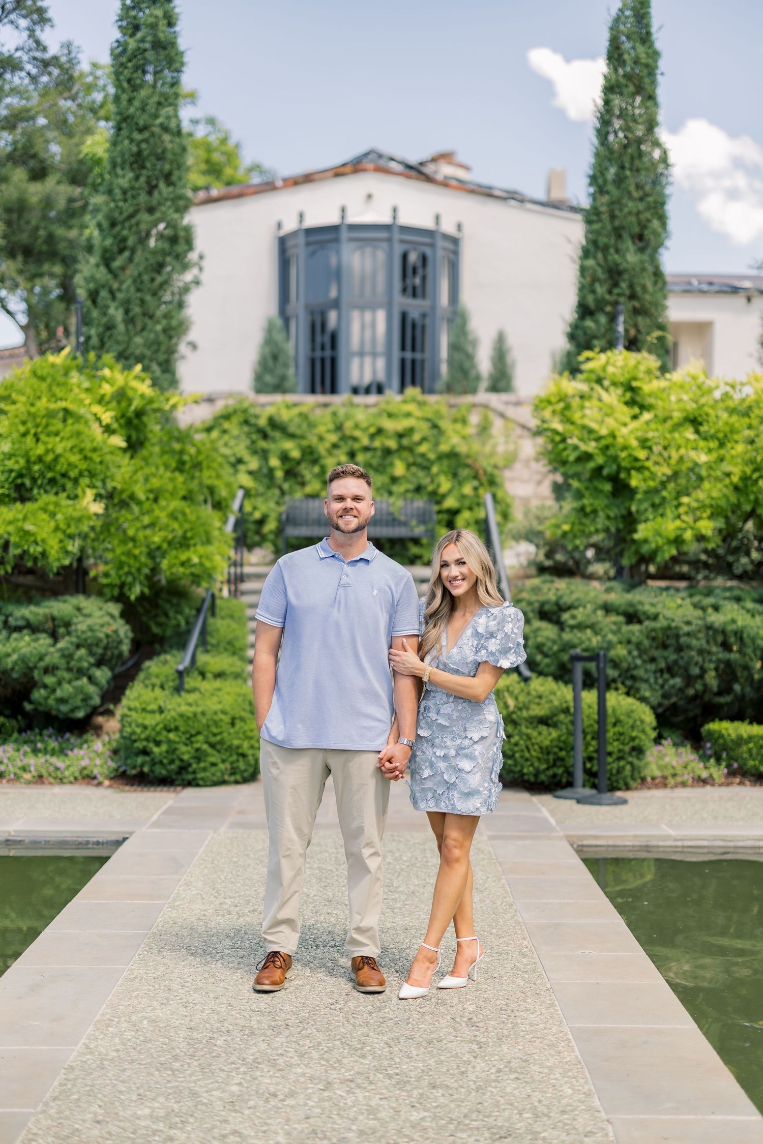 Couple holding hands smiling in front of elegant villa with lush green gardens and reflecting pool.